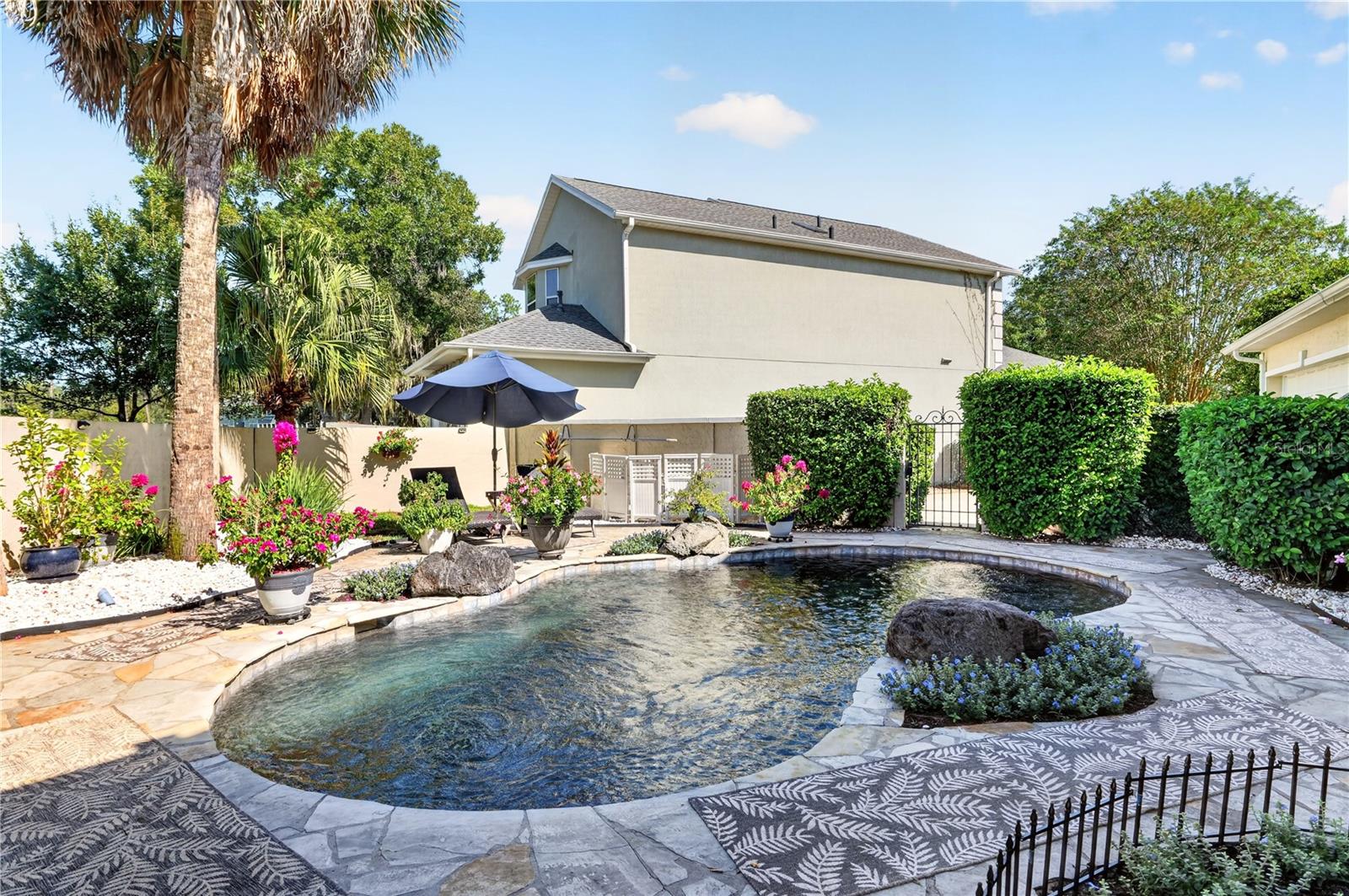 Backyard swimming pool with a curved stone edge, tropical plants, and a shaded patio umbrella.