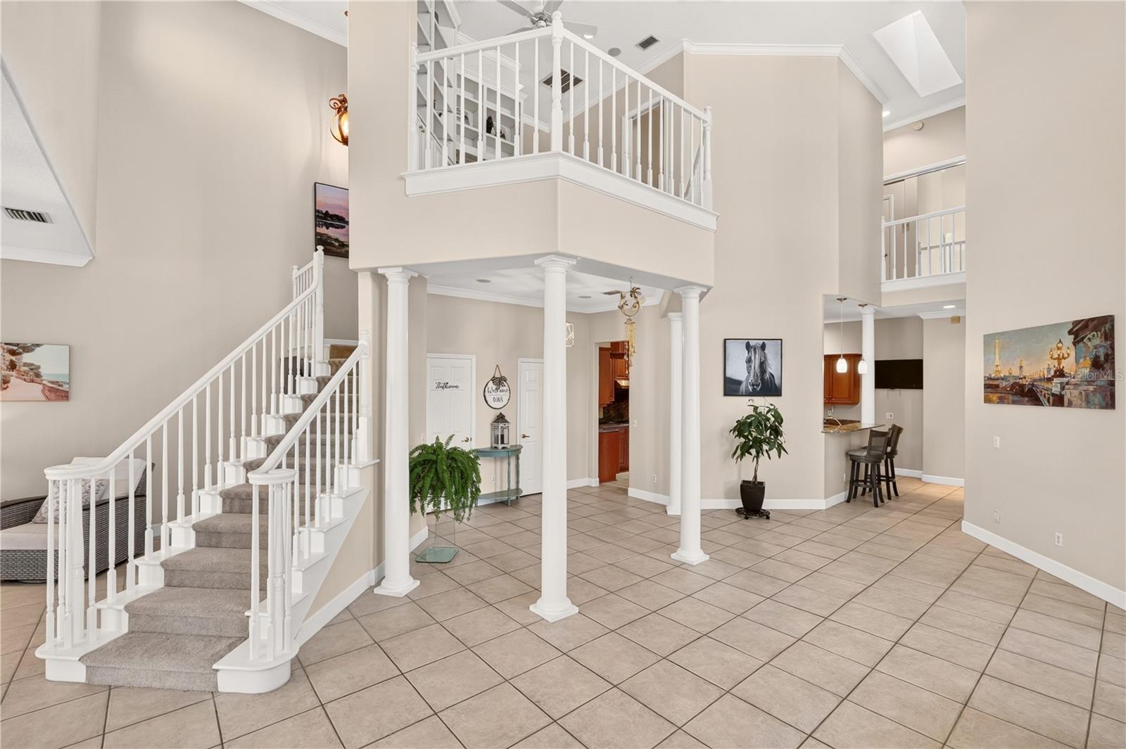 Two-story foyer with a carpeted staircase, white columns, tiled floor, and upstairs balcony.