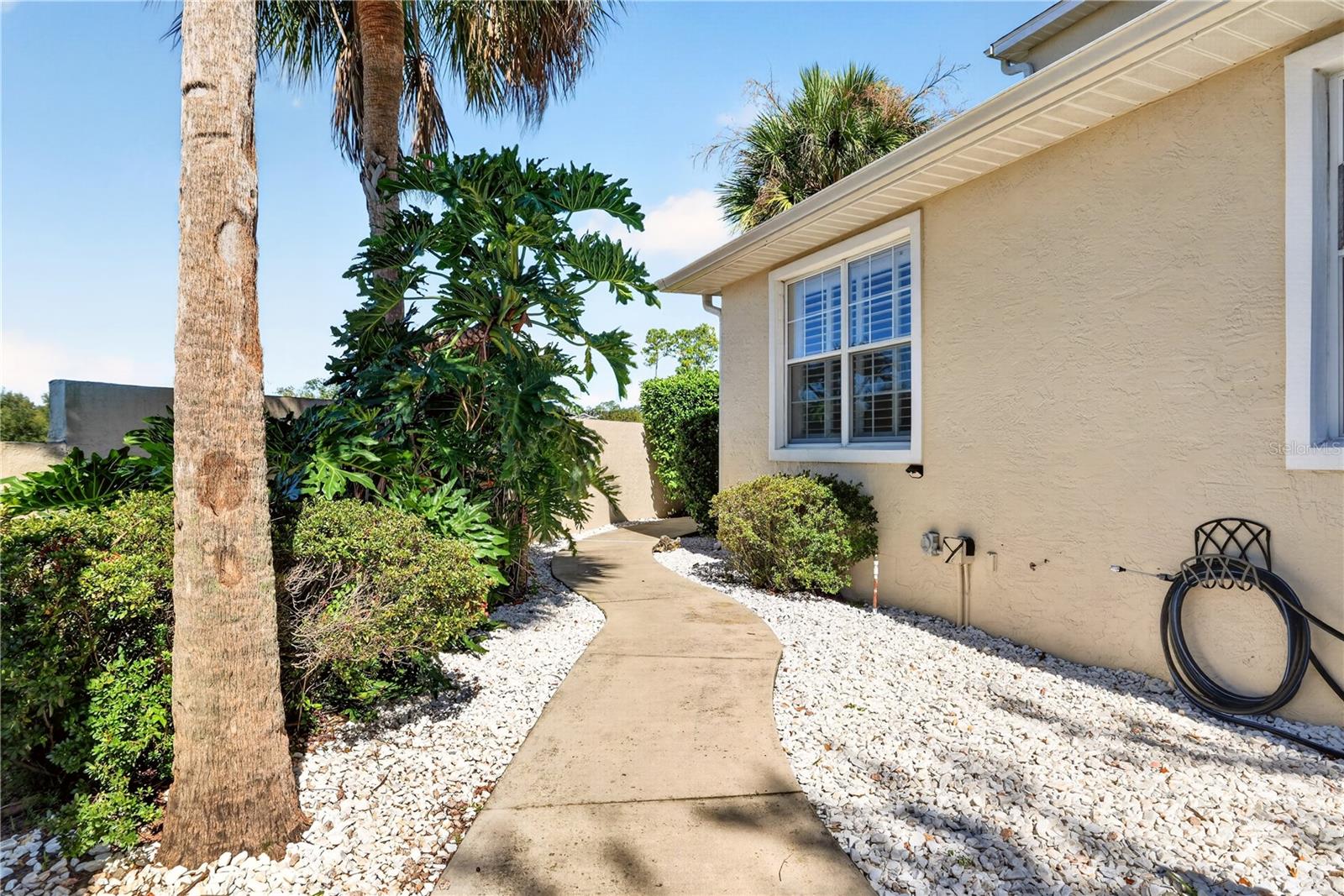 Beige stucco home beside a curved walkway bordered by white rocks, palms, and tropical shrubs.