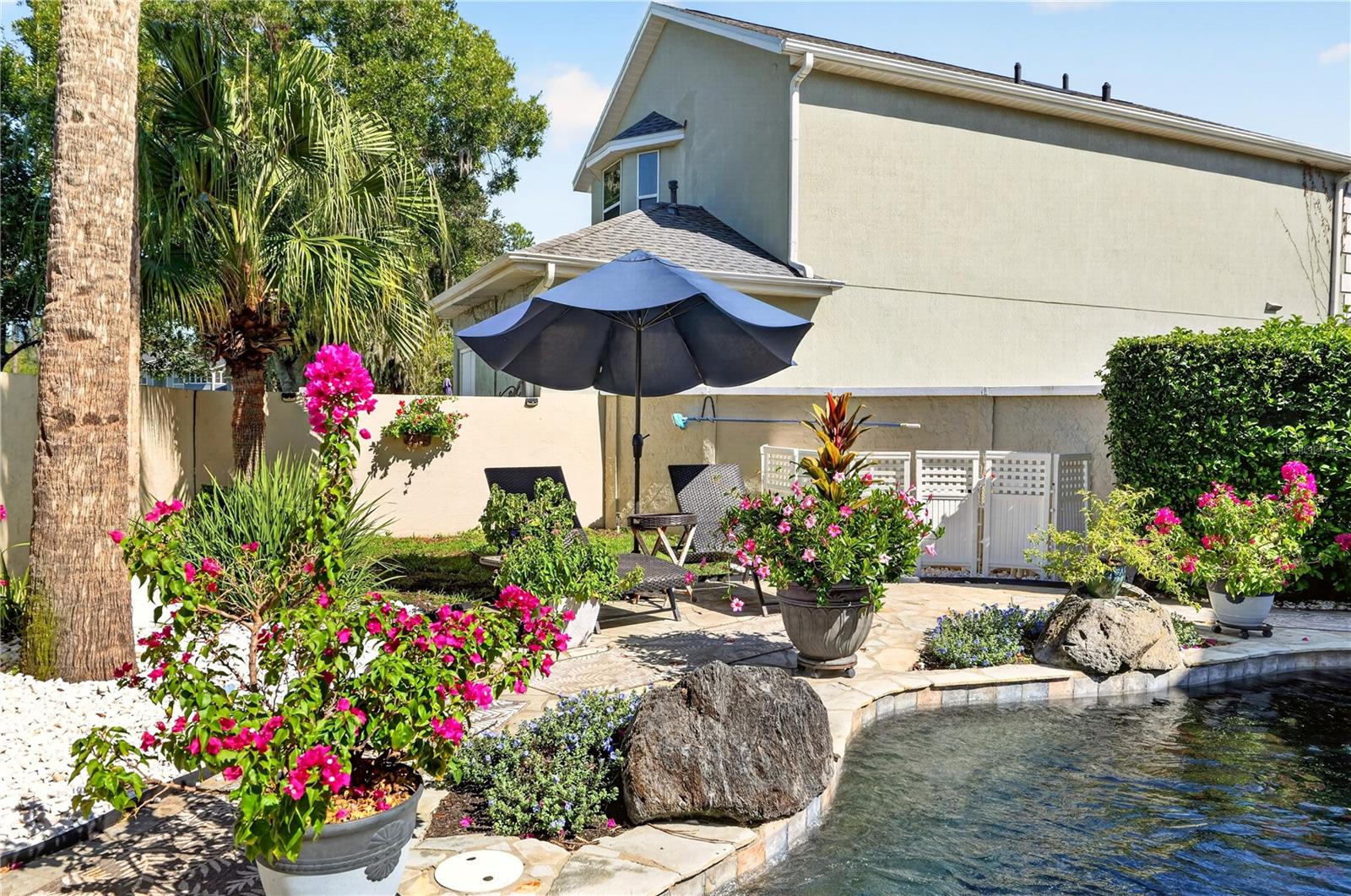Backyard pool beside a paved patio with an umbrella, seating, and flowering plants.