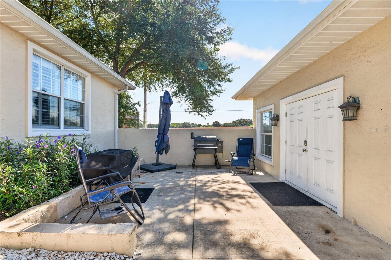 Paved patio courtyard with a barbecue grill, closed umbrella, folding chairs, and flowering planter.