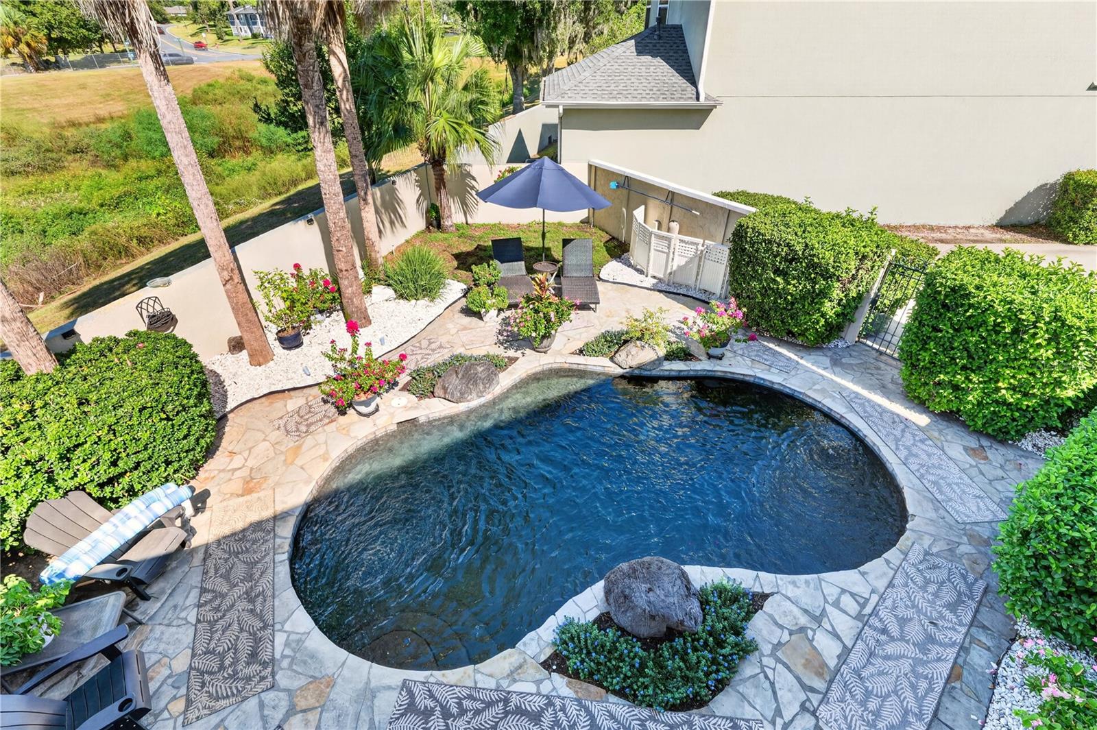 Curved backyard pool bordered by stone paving, tropical plants, and a shaded patio seating area.
