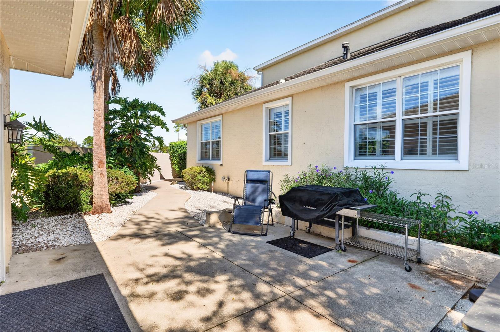 Side patio with a curved walkway, tropical landscaping, lounge chair, and covered grill.