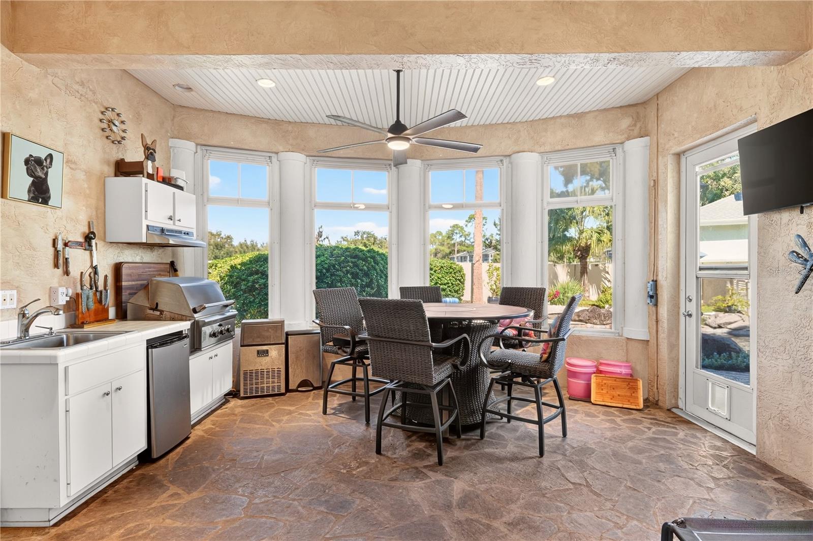 Sunroom with a wicker dining set, kitchenette, built-in grill, and garden views through large windows.