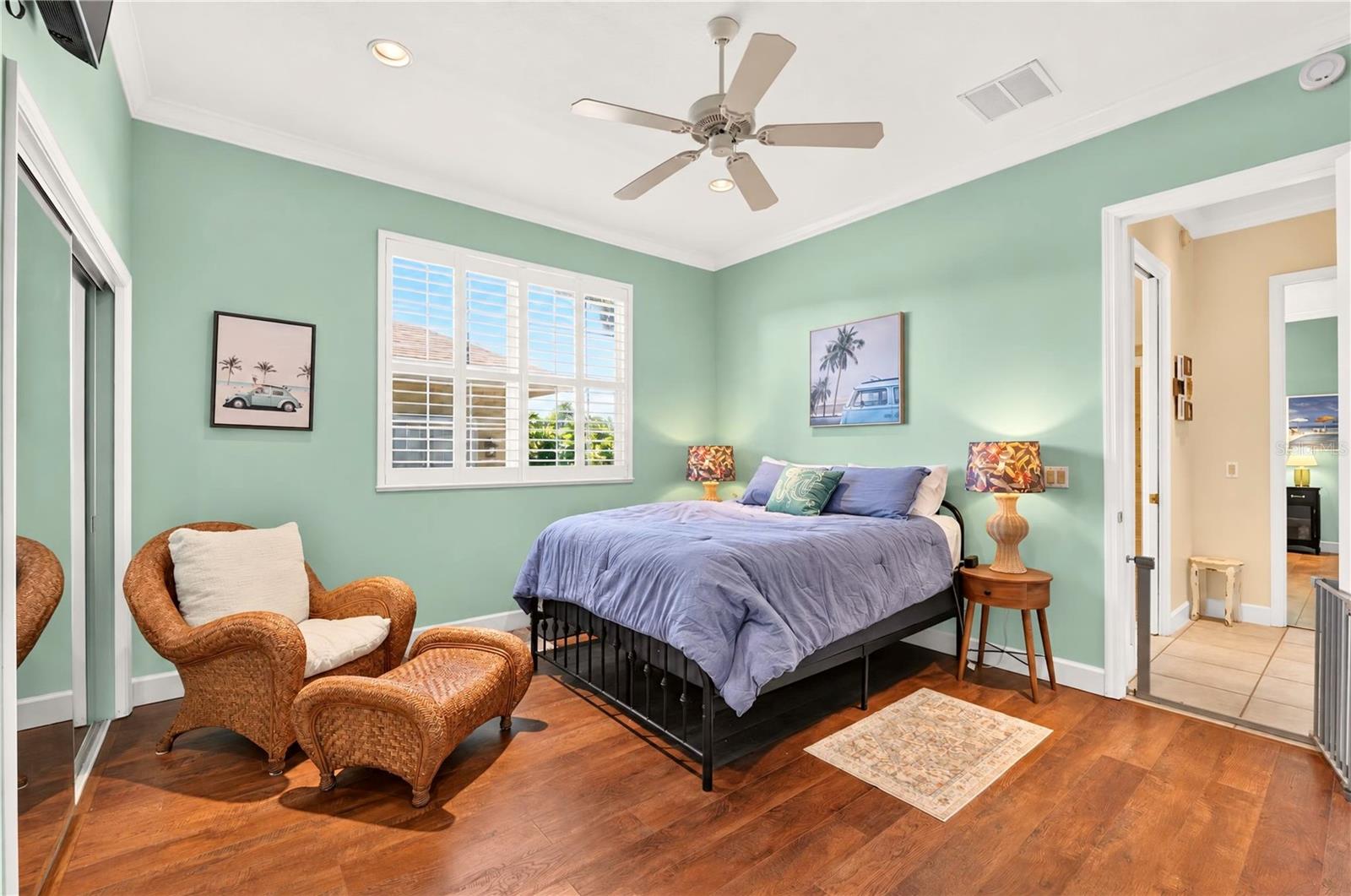 Mint-green bedroom with lavender bedding, plantation shutters, and a wicker armchair and ottoman.