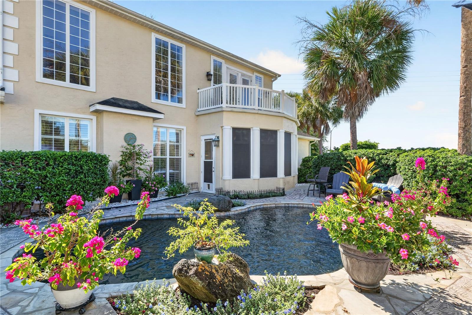 Two-story beige home beside a courtyard pool, with a balcony, palm trees, and flowering plants.