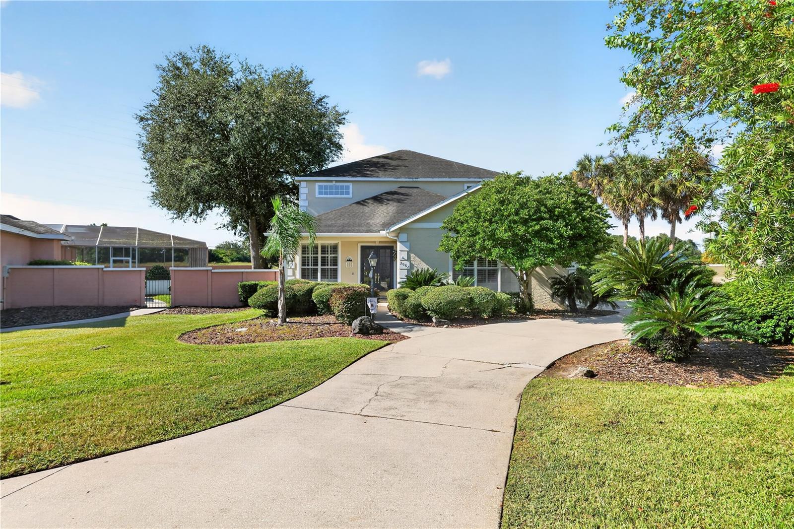Two-story gray-green home behind a curved driveway, with a landscaped lawn and palm trees.