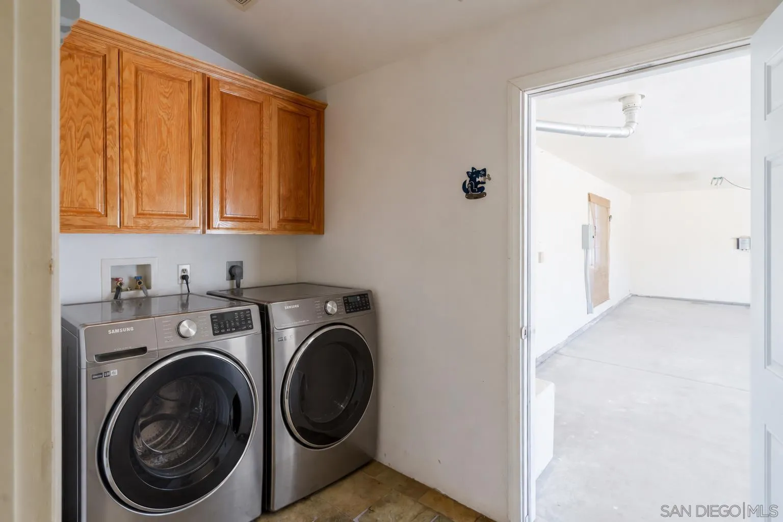 Laundry room with front-loading washer and dryer beneath wooden cabinets, opening to a bright adjoining room.