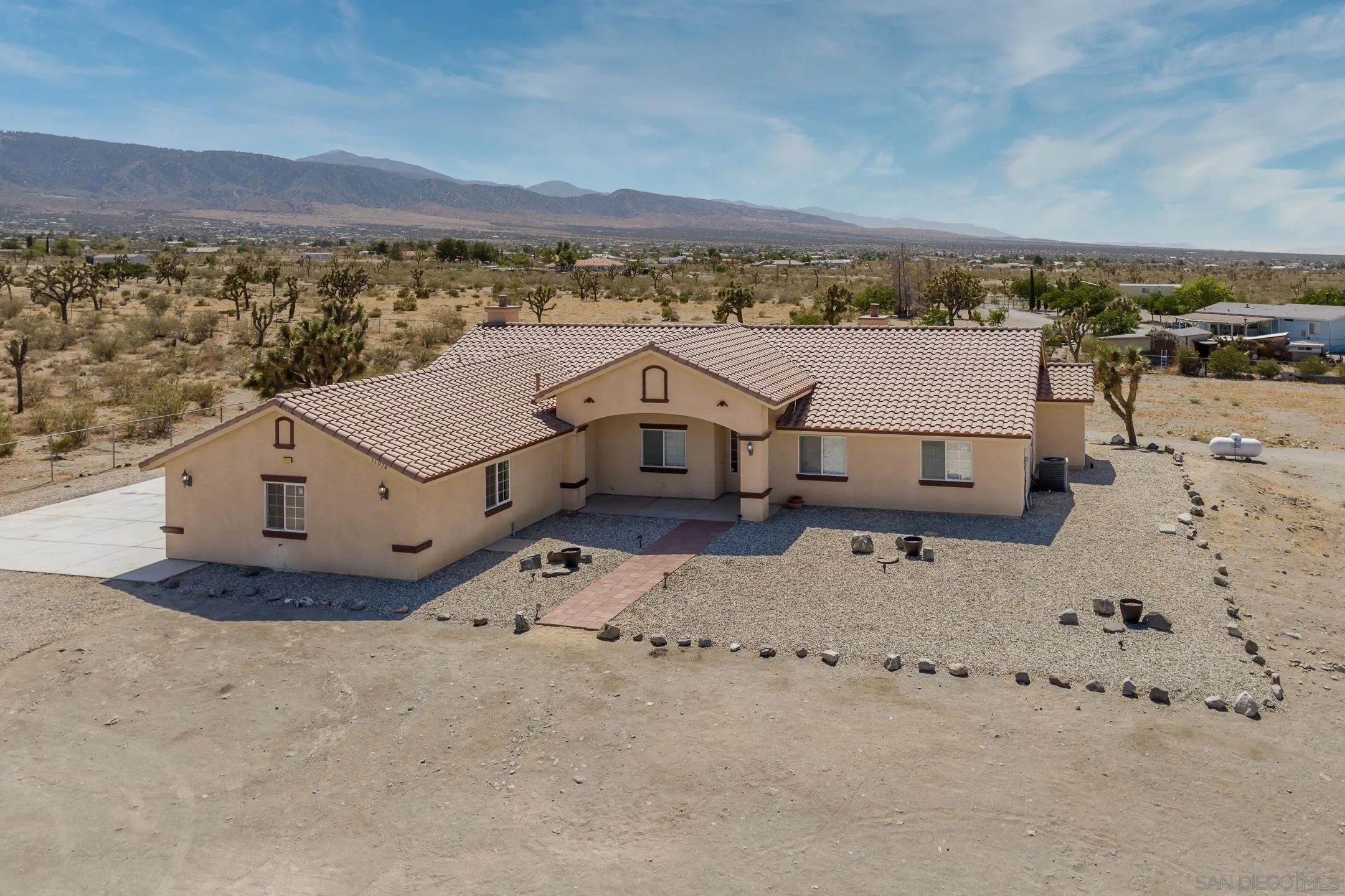 Aerial view of a tan, tile-roofed home on a gravel lot with desert mountains beyond.