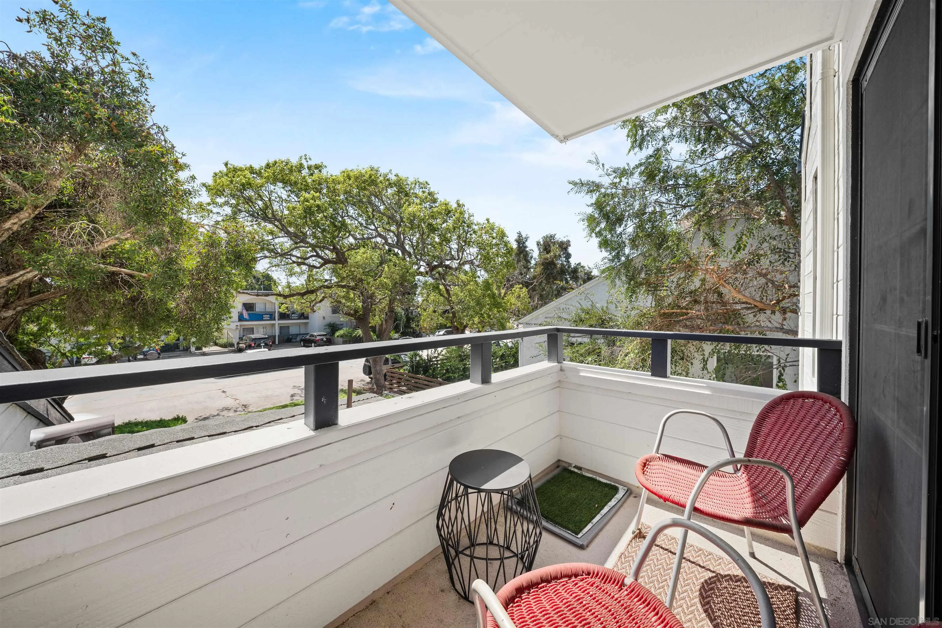 Apartment balcony with red woven chairs and a small table overlooking a tree-lined street.