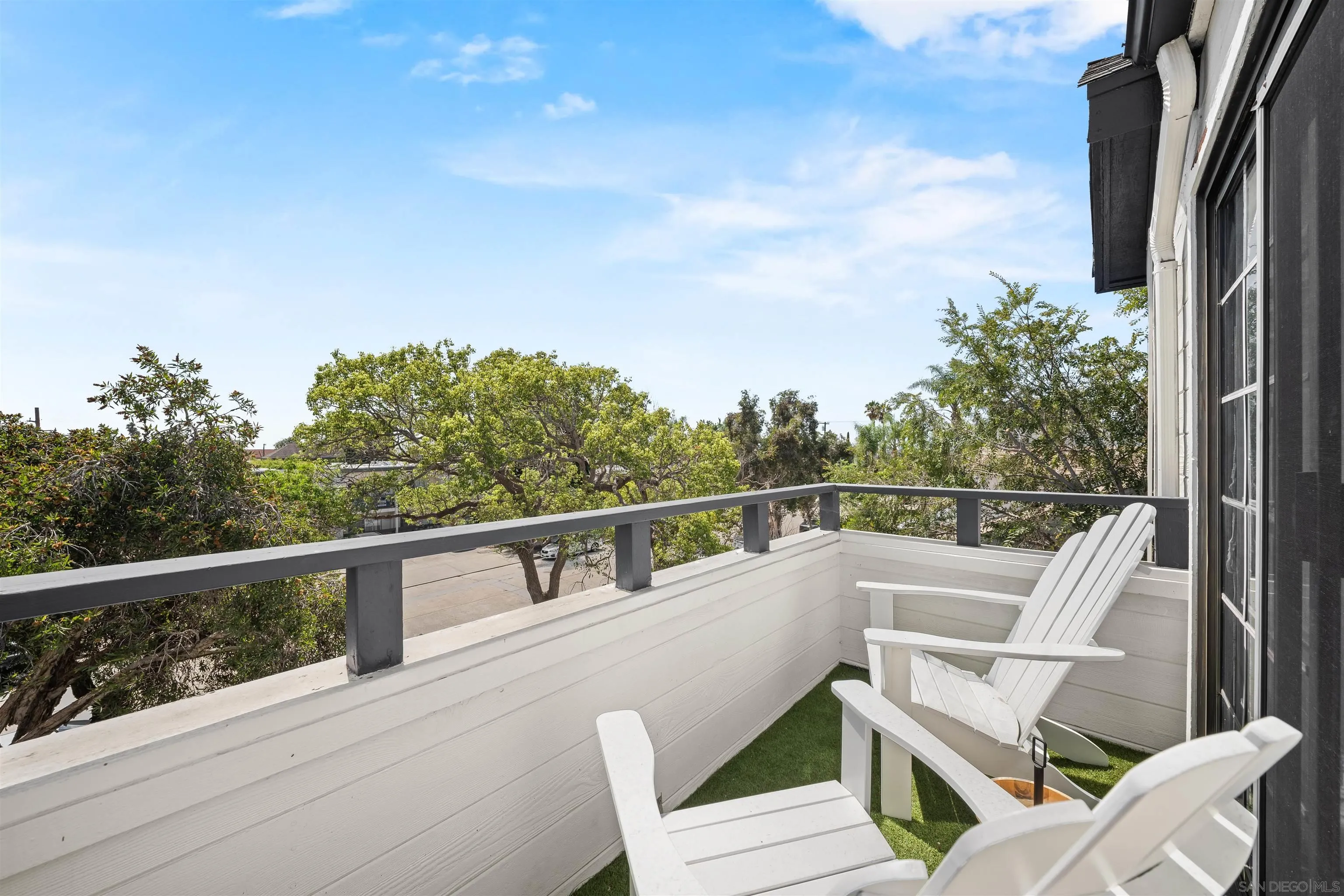 Elevated deck with white Adirondack chairs, turf flooring, and leafy trees beyond the railing.