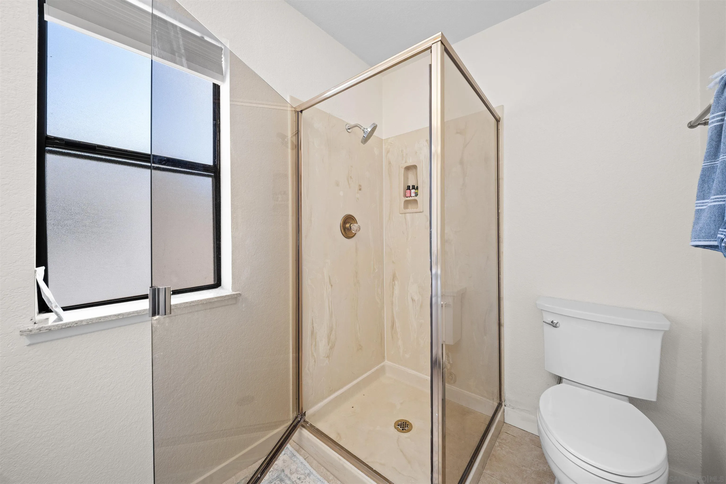 Bathroom with a corner glass shower, beige stone-look walls, a frosted window, and a white toilet.
