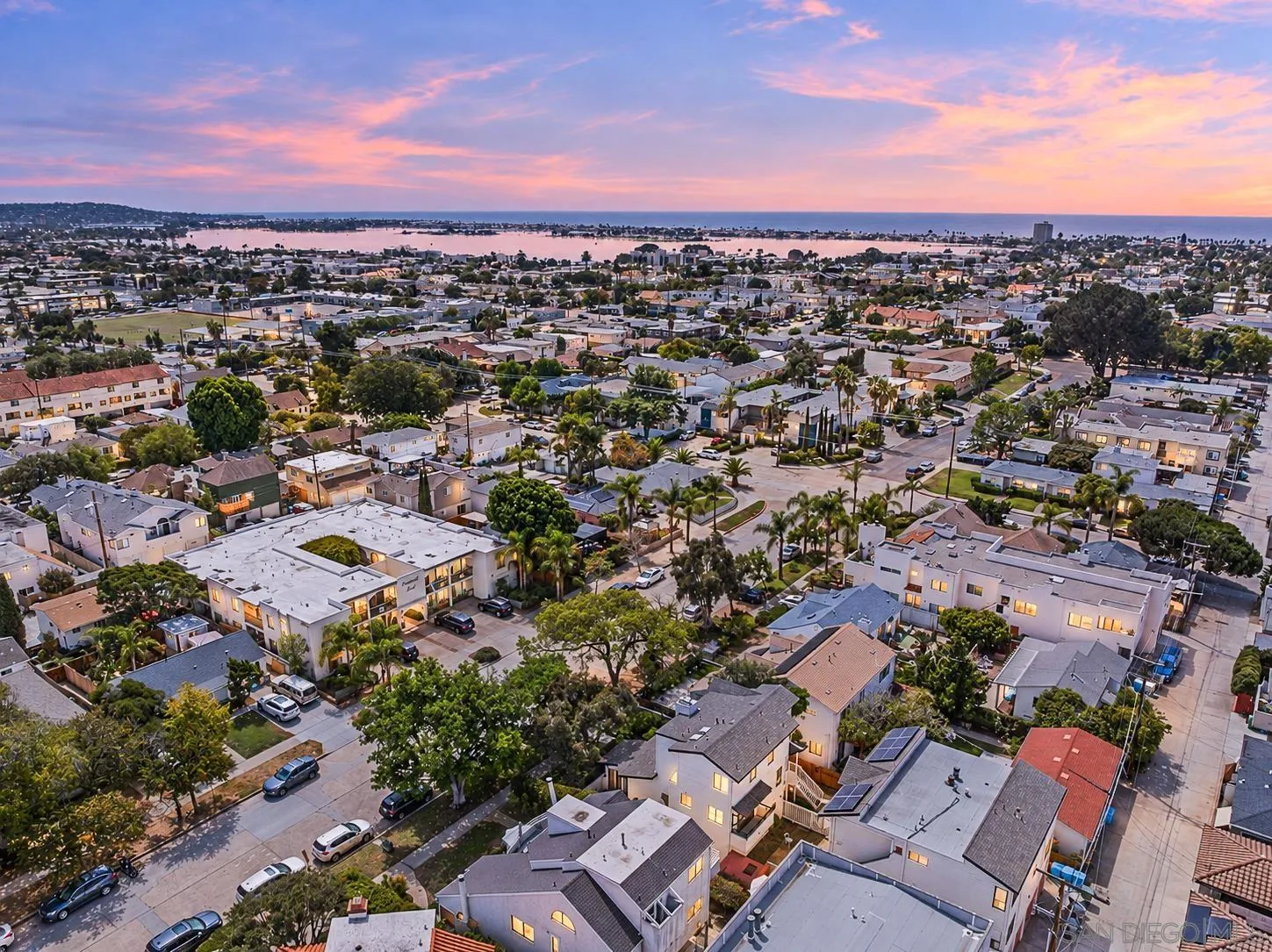 Aerial view of coastal homes and palm trees with the bay and ocean beyond at sunset.