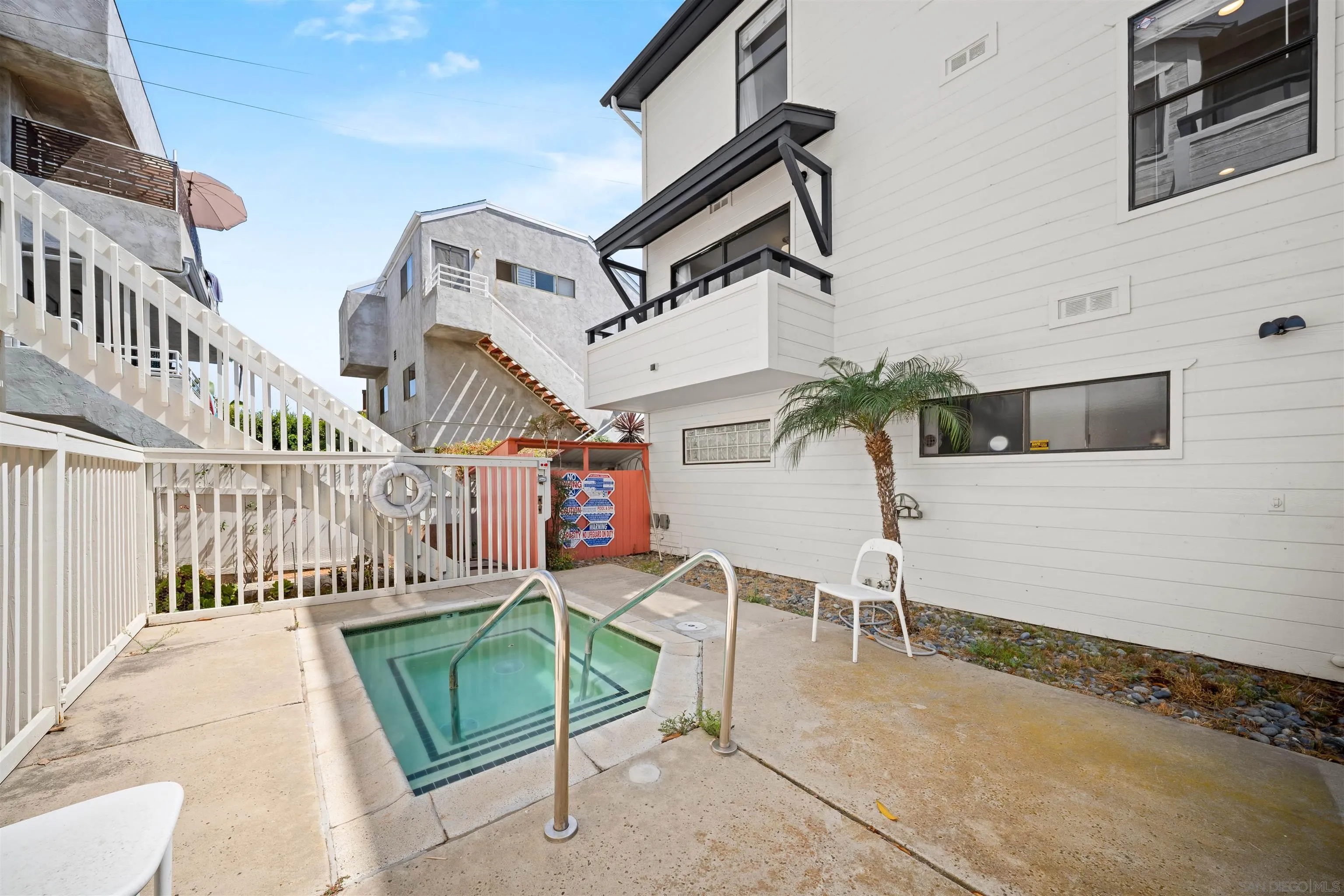 Small courtyard pool beside a white multi-story building, enclosed by railings with a palm tree nearby.
