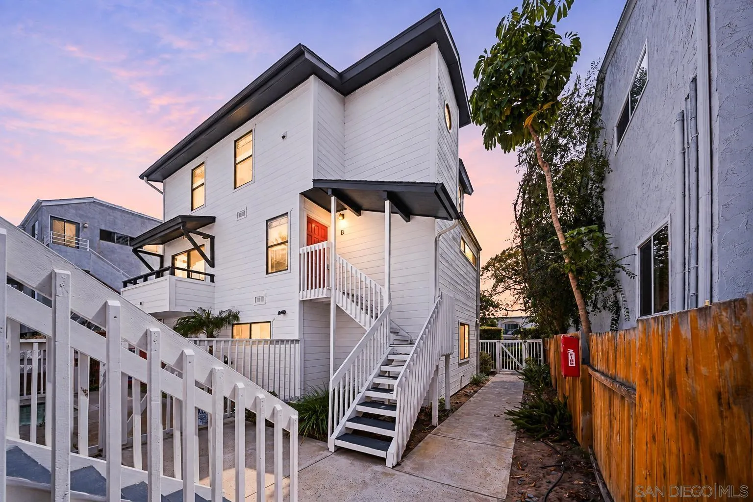 White two-story home with a red Unit B door, exterior stairs, and a narrow fenced walkway at dusk.