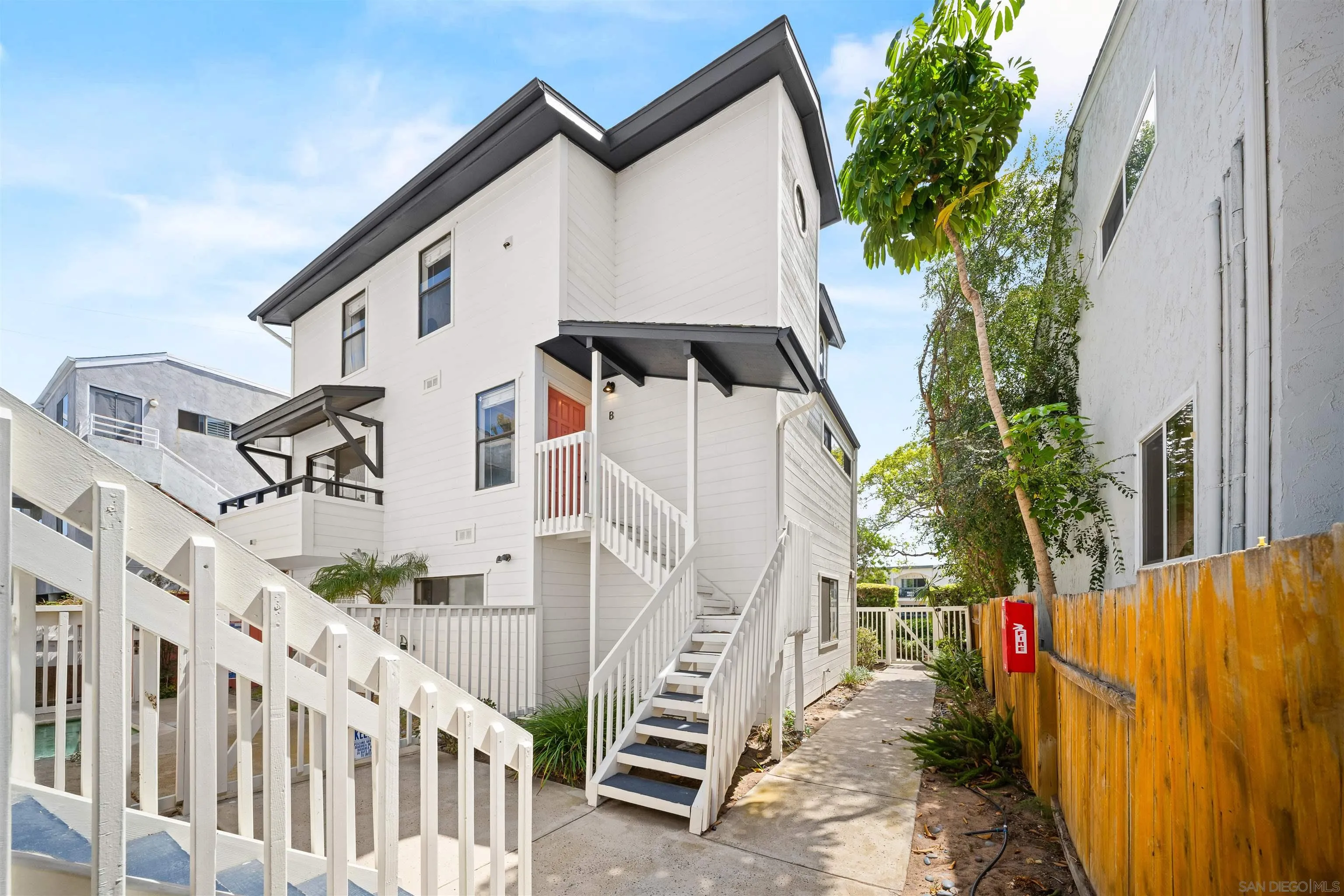White multi-story residence with a red door marked B, exterior stairs, and a tree-lined side walkway.