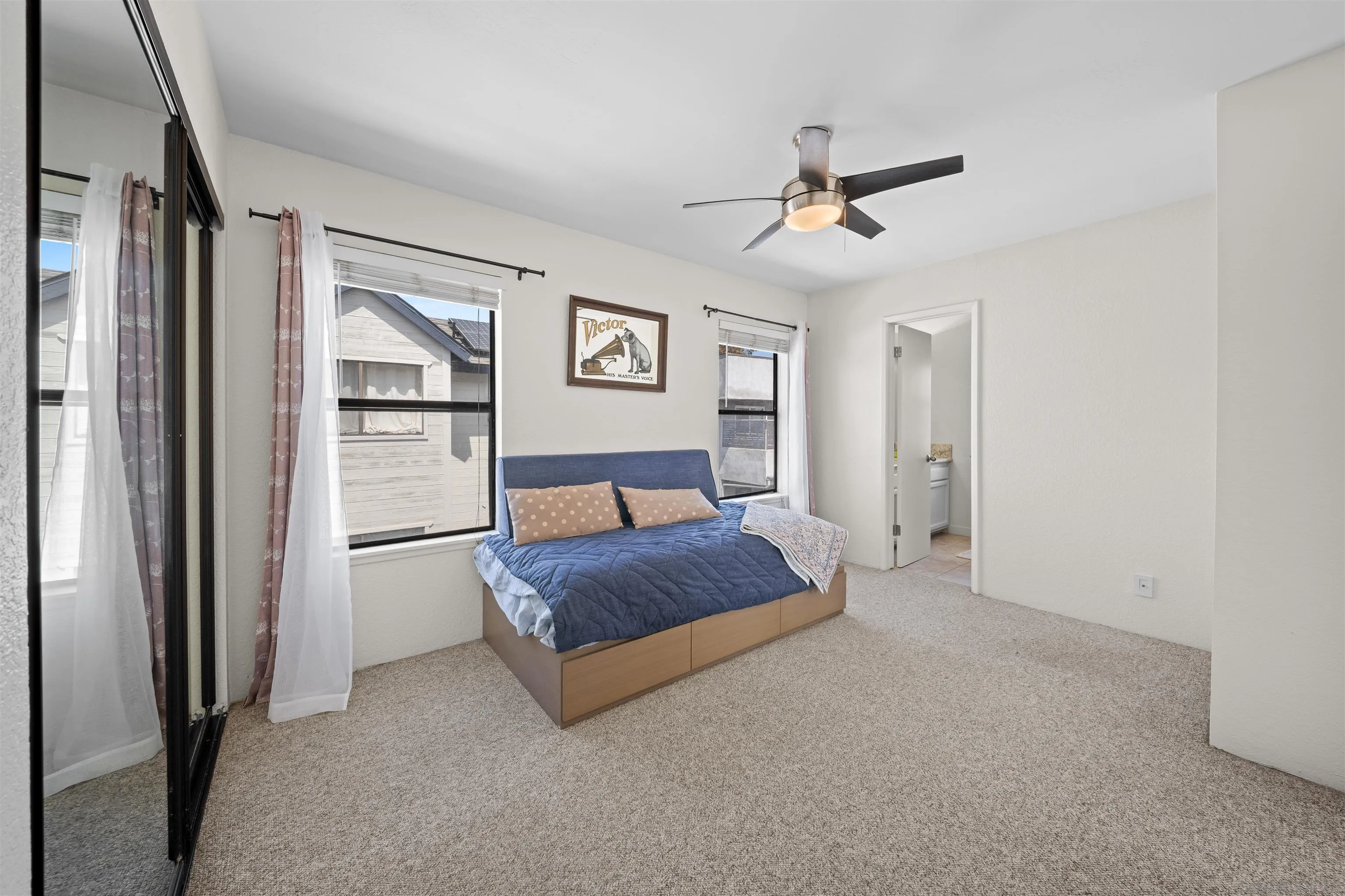 Bedroom with a blue-quilted bed, mirrored closet doors, curtained windows, and ceiling fan.