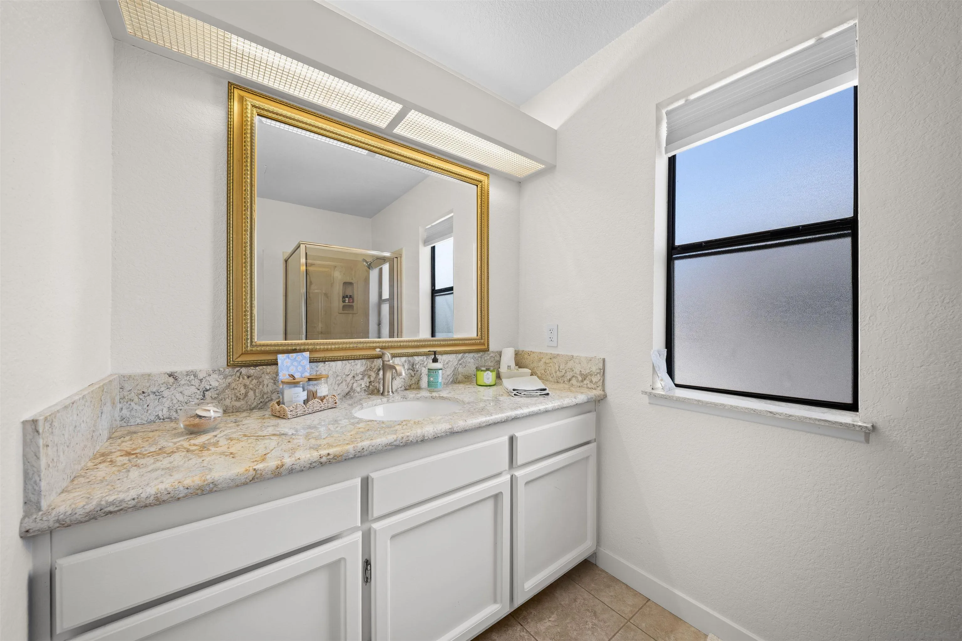 Bathroom vanity with granite counter, white cabinetry, ornate gold-framed mirror, and a window.