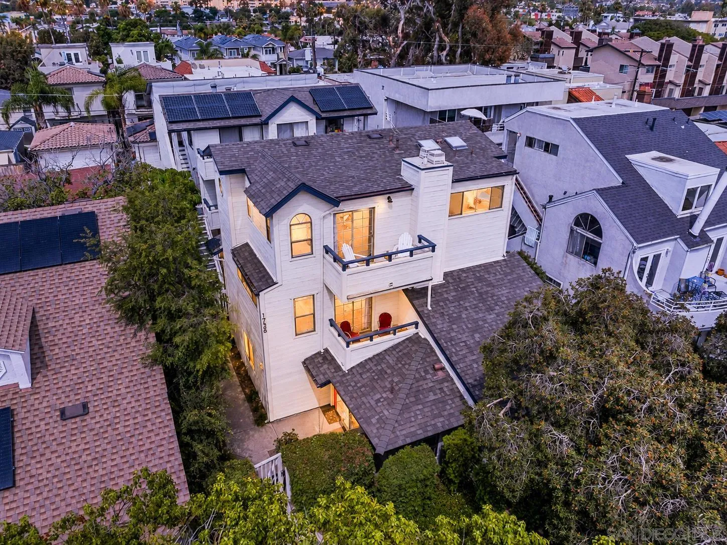 Aerial view of a white multi-story home with two balconies and warmly lit windows among neighboring homes.