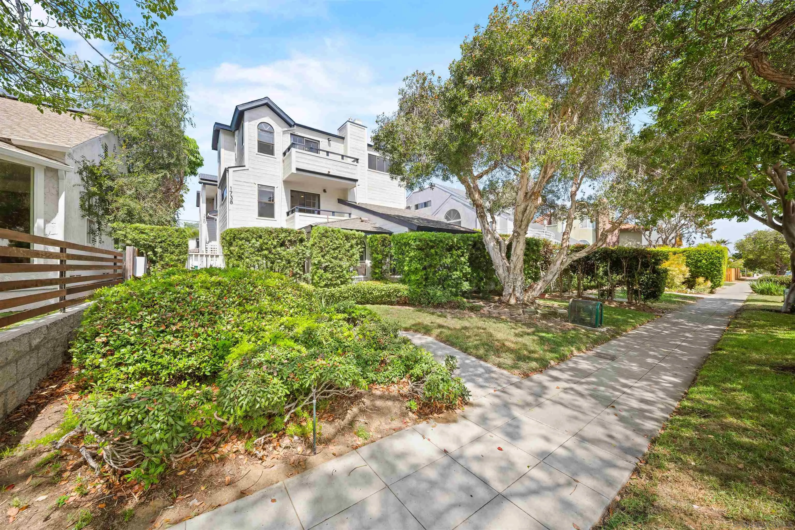 White multi-story residence with balconies behind mature trees and landscaped hedges.