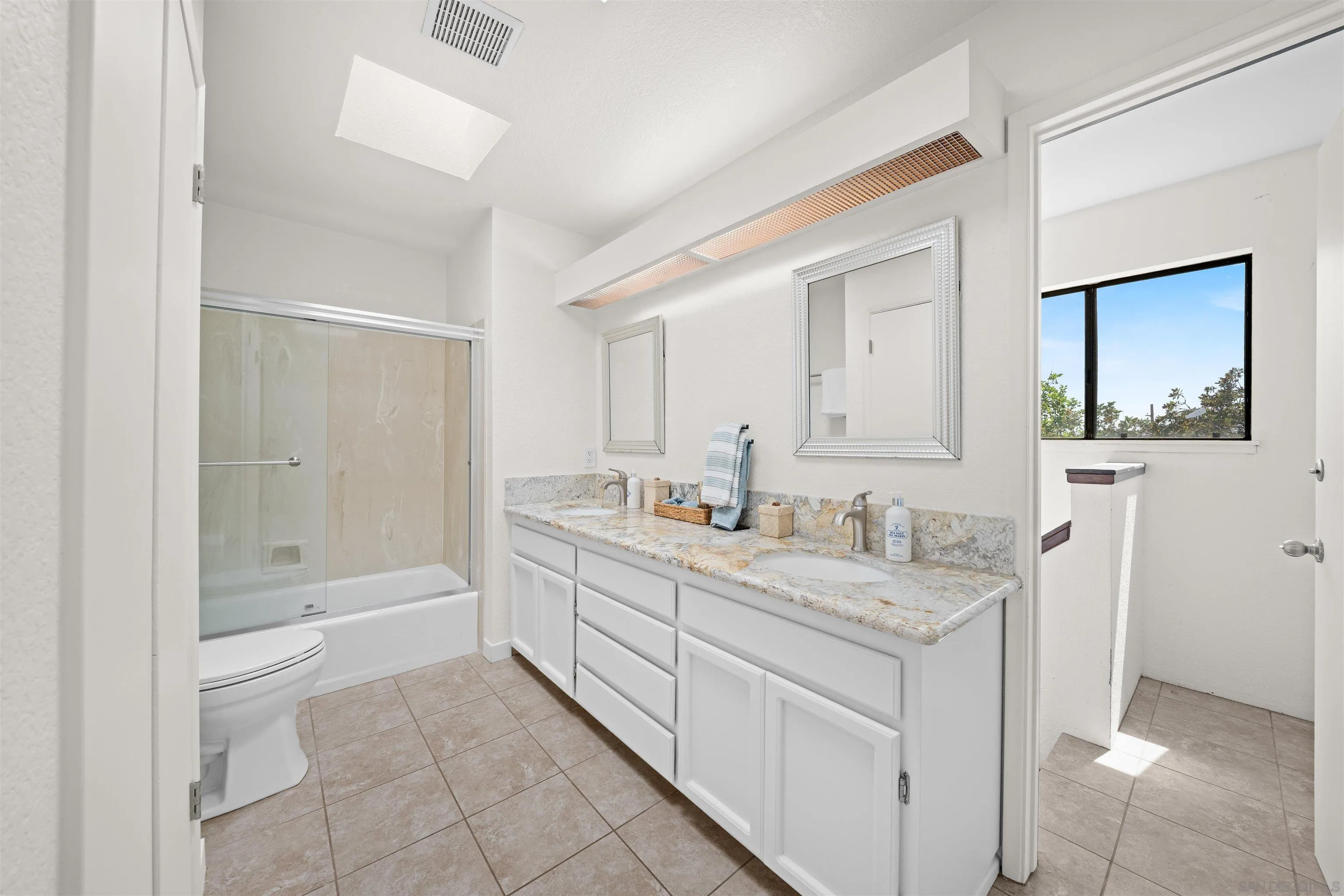 Bright bathroom with a double-sink vanity, marbled countertop, tiled tub-shower, and window overlooking greenery.
