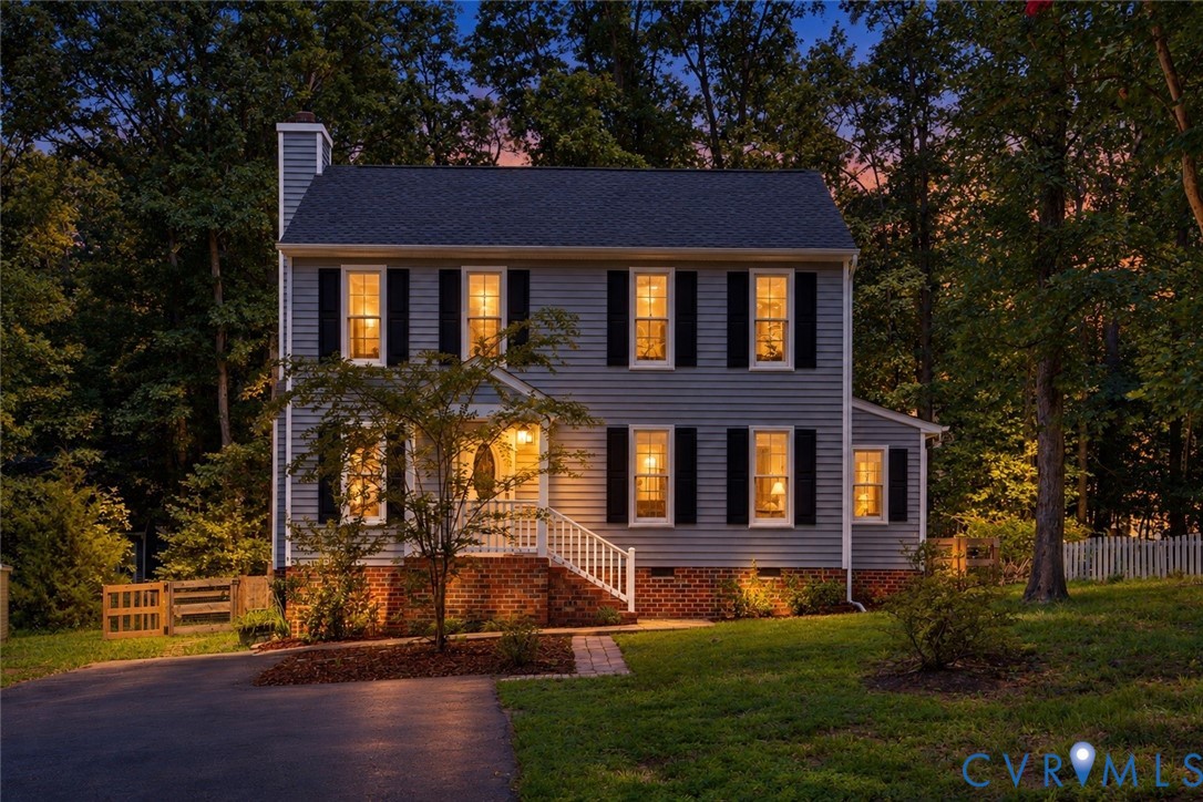 Two-story gray house with a brick foundation and warmly lit windows among trees at dusk.
