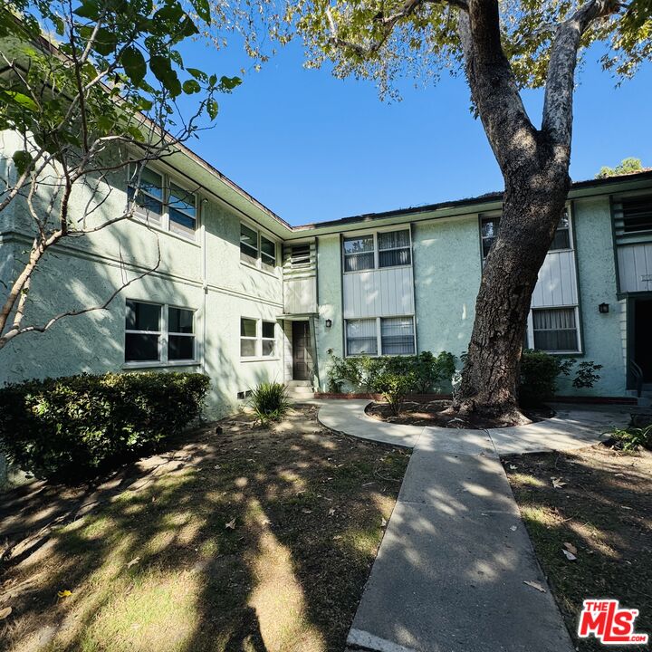 Two-story pale green apartment building behind a courtyard walkway and a large mature tree.