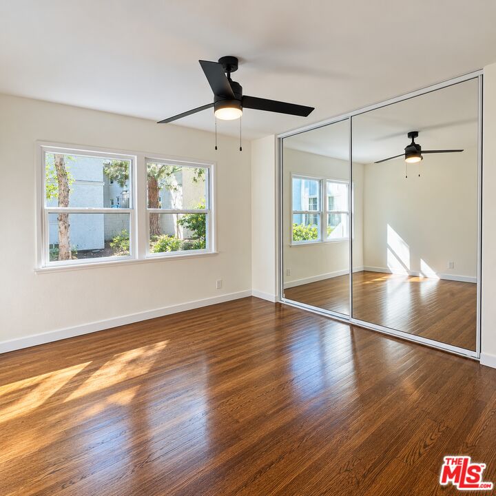Empty bedroom with polished wood floors, mirrored closet doors, two windows, and a ceiling fan.
