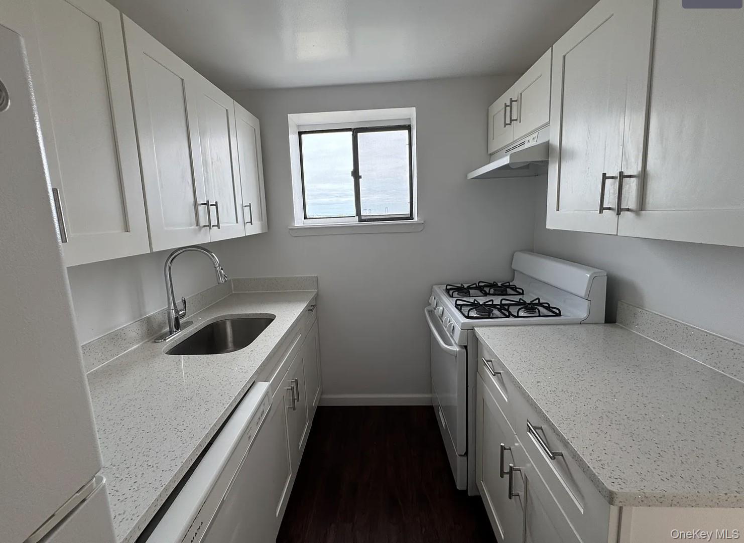 Galley kitchen with white cabinets, speckled countertops, a gas range, and a window at the far end.