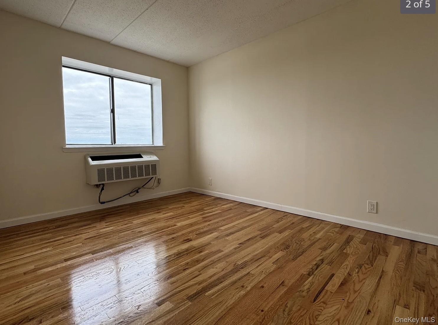 Empty apartment room with polished hardwood floors, a window, and a through-wall air conditioner.