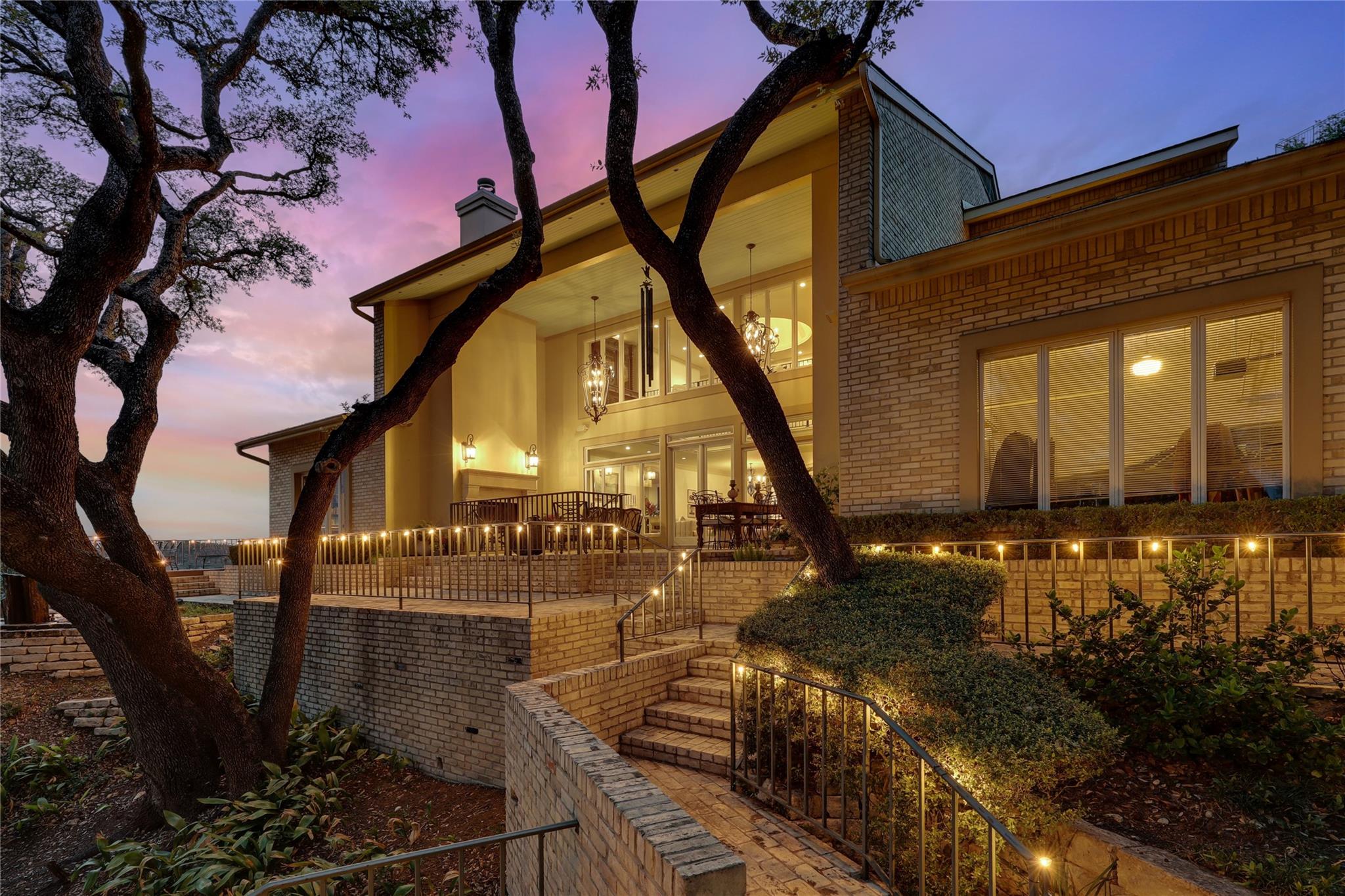 Two-story brick home at dusk with a warmly lit patio, brick steps, and mature trees.