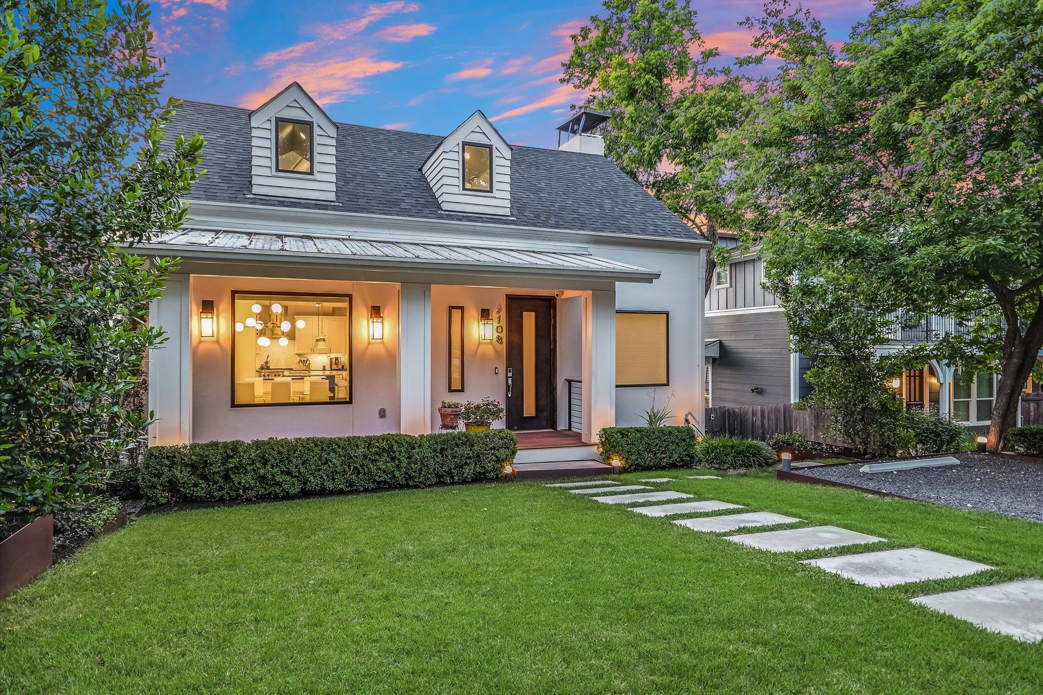 White two-story home with dormer windows, a warmly lit entry, and stepping stones across the lawn at dusk.