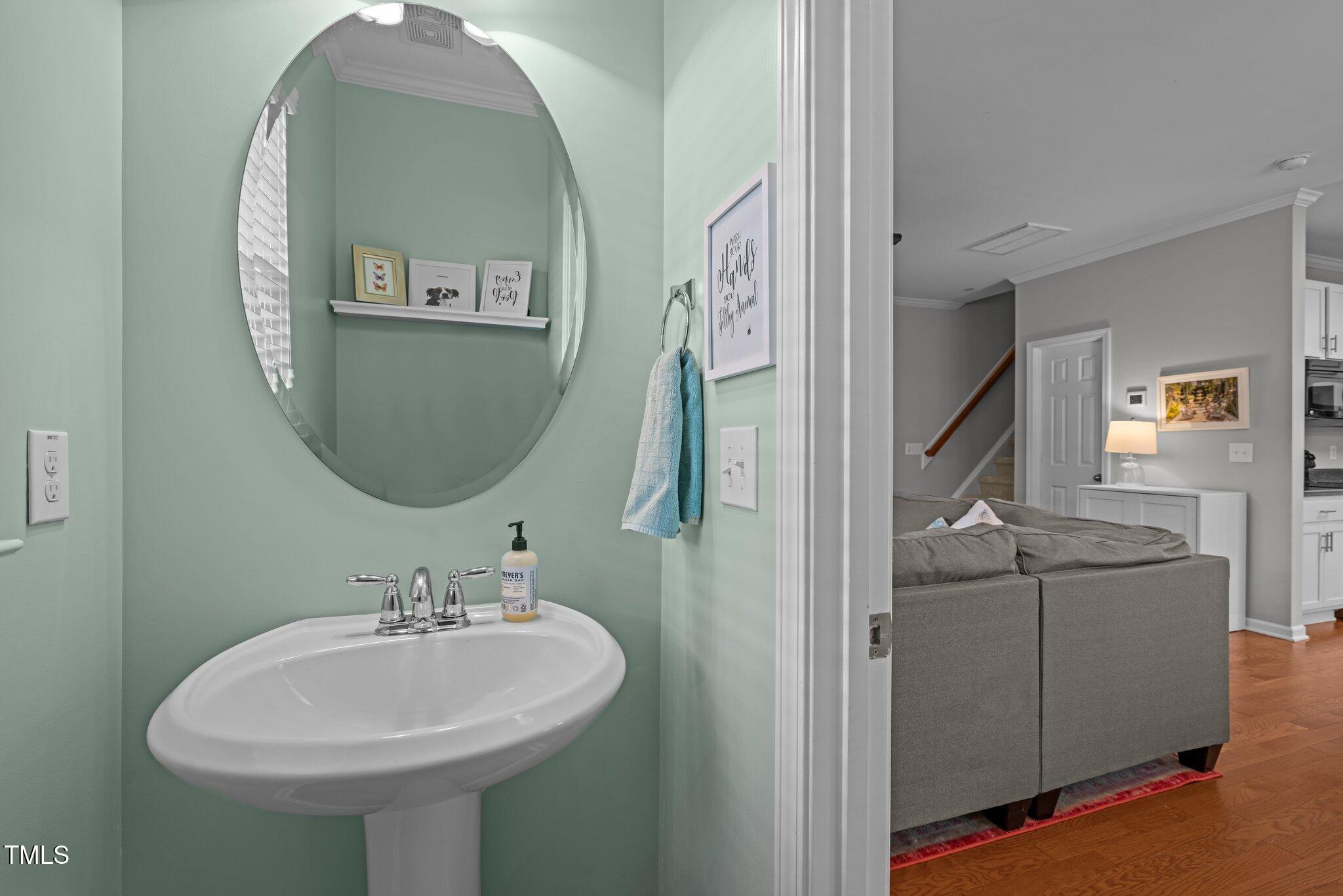 Mint-green bathroom with oval mirror and pedestal sink opening onto a living room with a gray sofa.