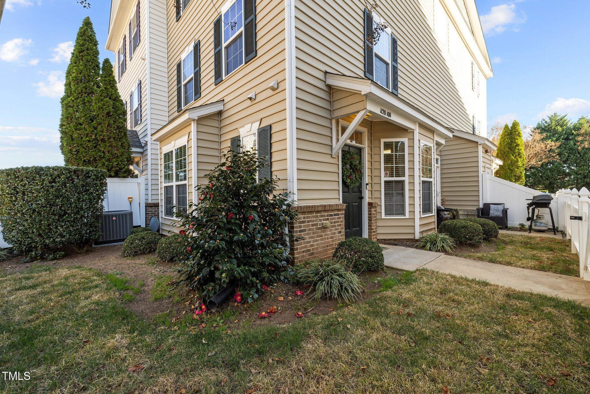 Beige-sided residence with a covered entrance, landscaped shrubs, white fencing, and a small outdoor seating area.