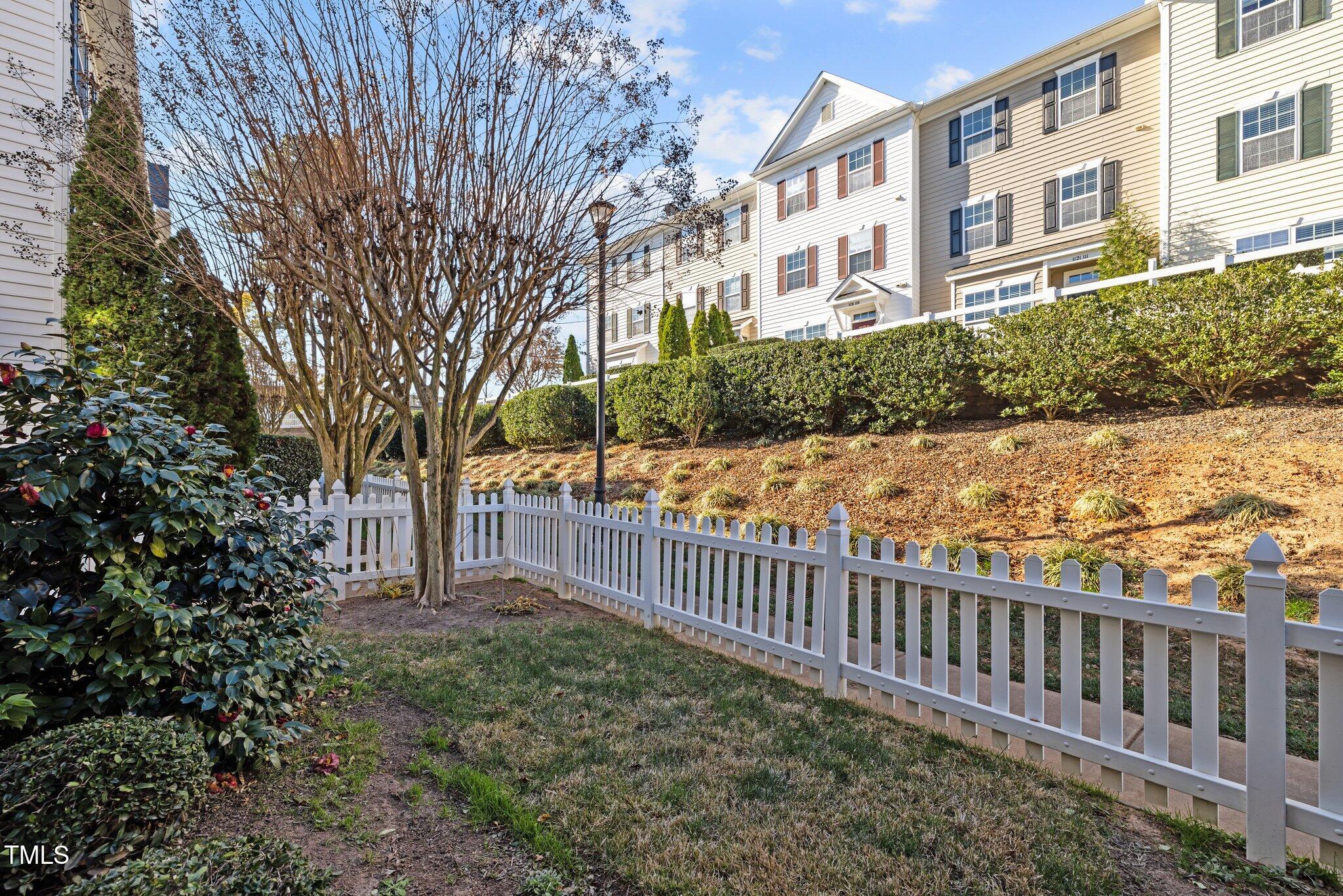 Townhouse exterior beyond a white picket fence, with a leafless tree and flowering shrubs in the yard.