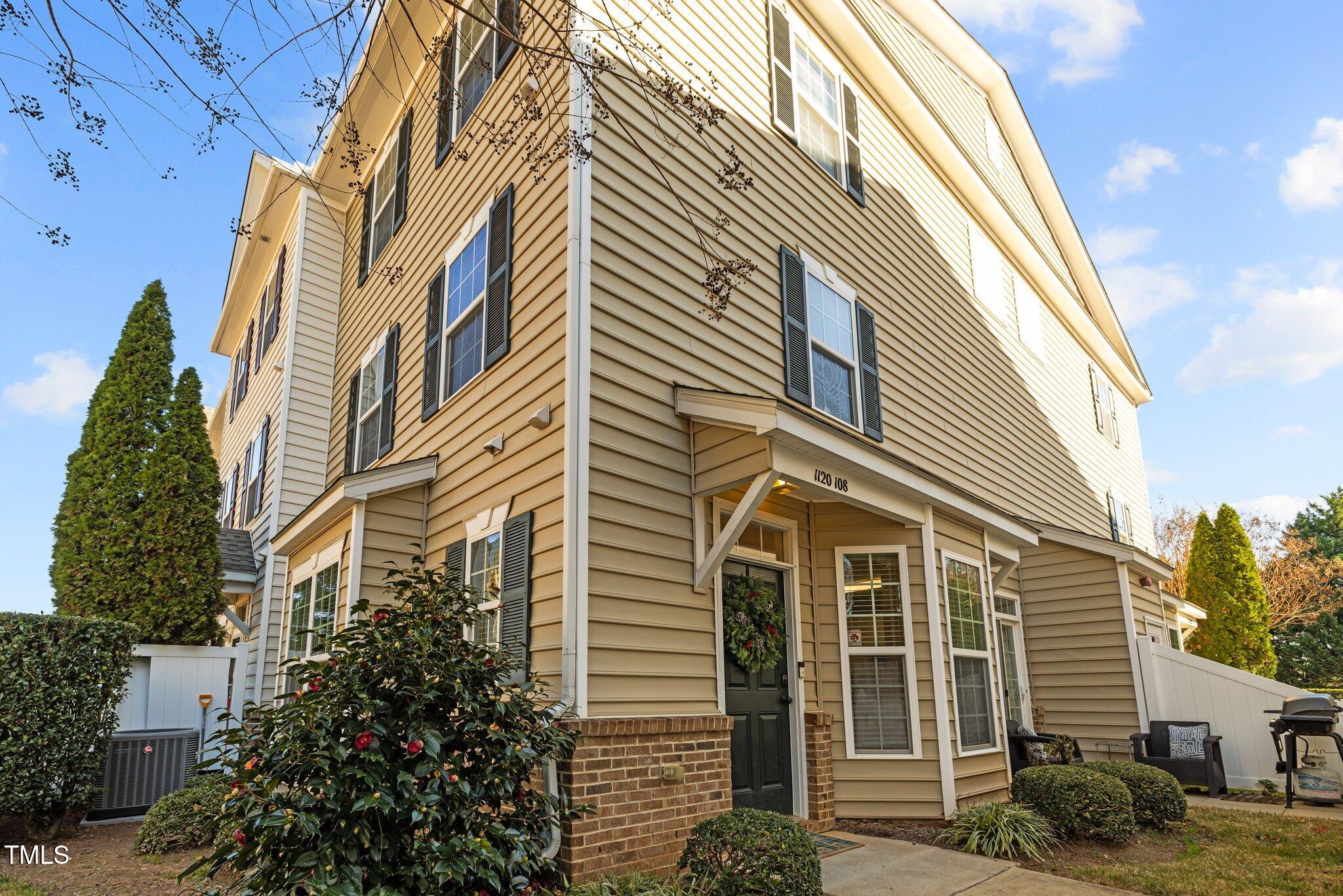 Beige townhouse exterior with black shutters, a covered entrance, landscaped shrubs, and a small fenced patio.