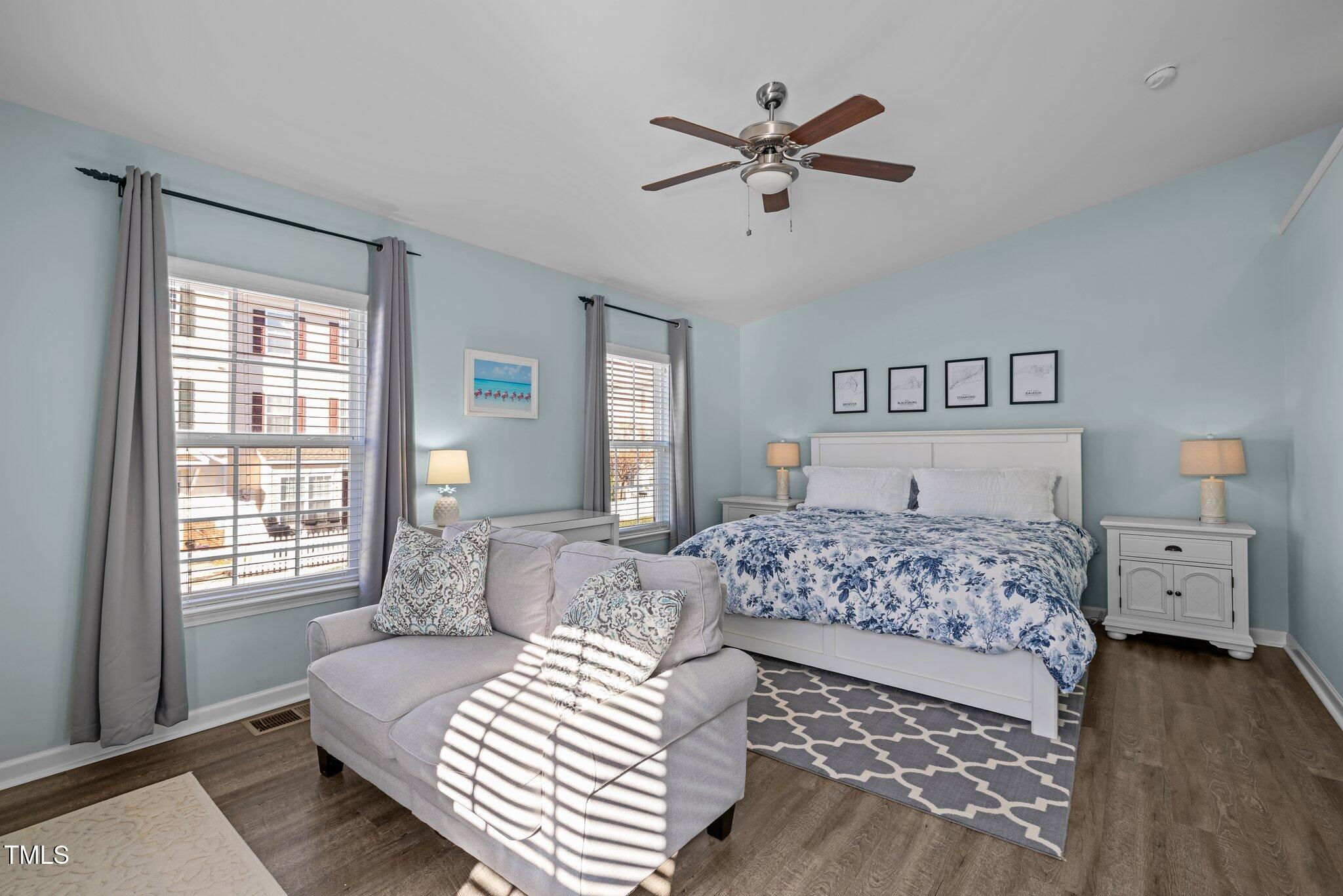 Bedroom with a white bed in blue floral bedding, gray chaise, and sunlight through two windows.