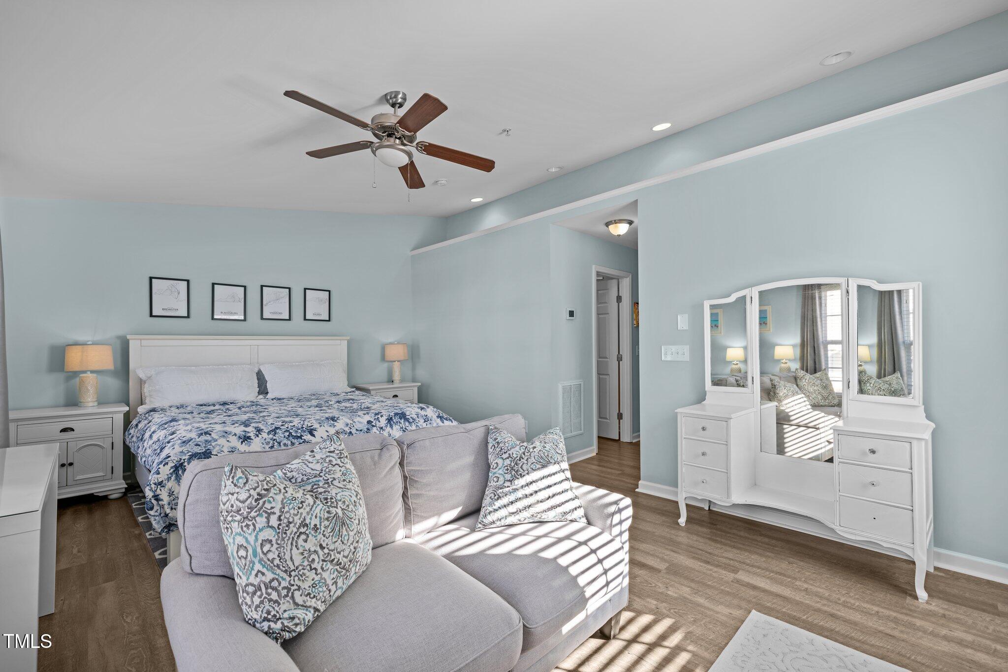 Bedroom with blue floral bedding, mirrored white dresser, and gray sofa at the foot of the bed.