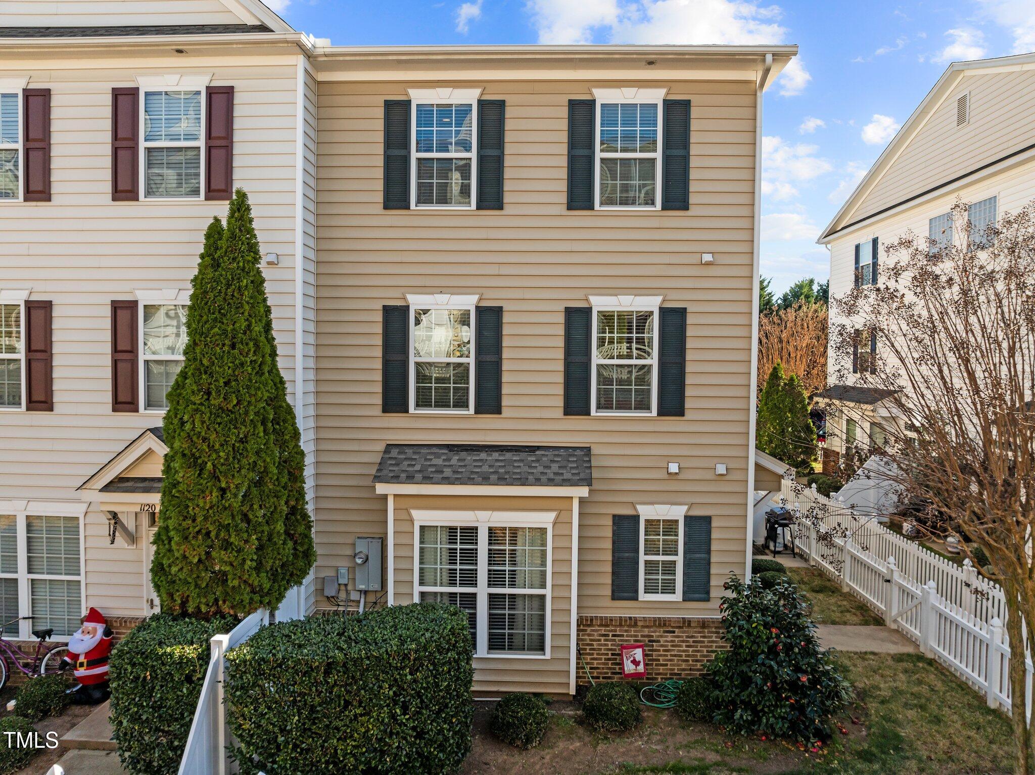 Beige townhome with dark shutters, brick foundation, and a fenced yard with a tall evergreen.