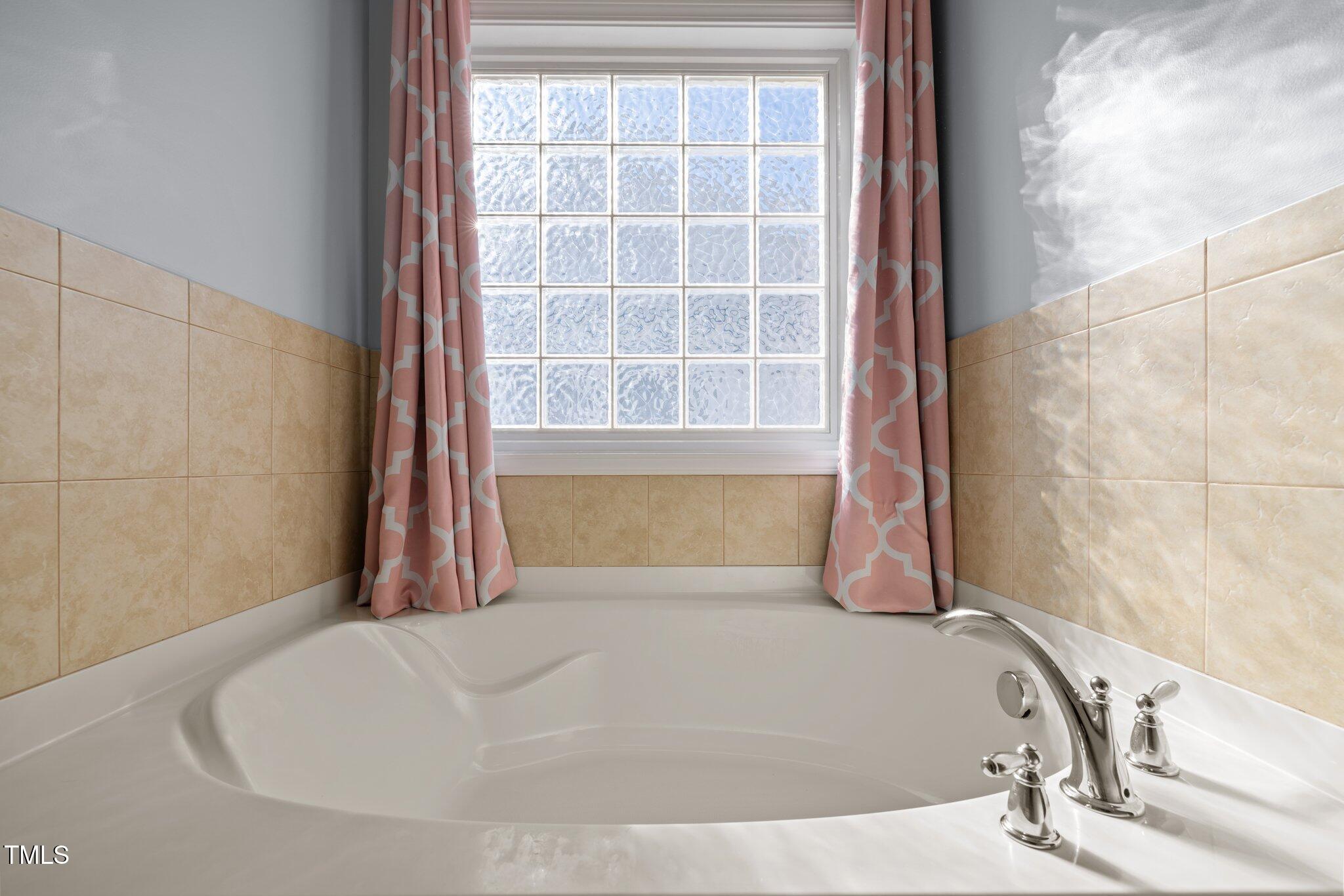 Corner soaking tub with chrome fixtures beneath a textured-glass window and pink curtains.