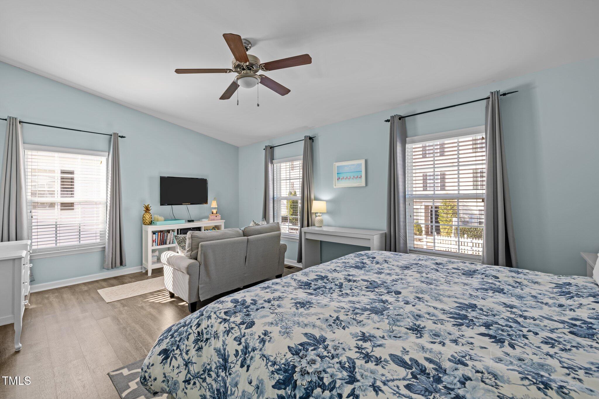 Furnished bedroom with a blue floral bed, gray loveseat, three curtained windows, and ceiling fan.