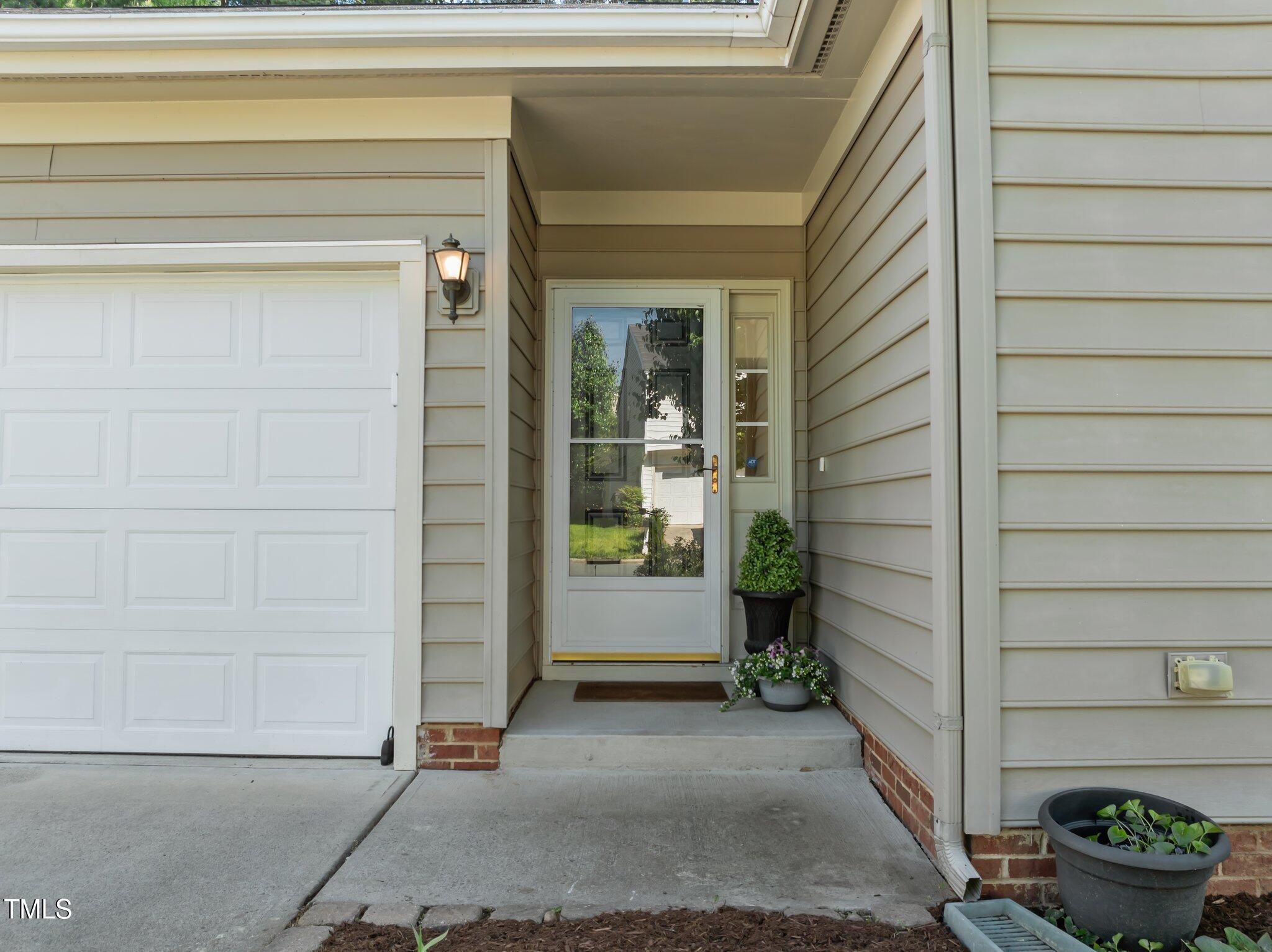 Covered front entry with a glass storm door, potted plants, and an adjacent white garage door.