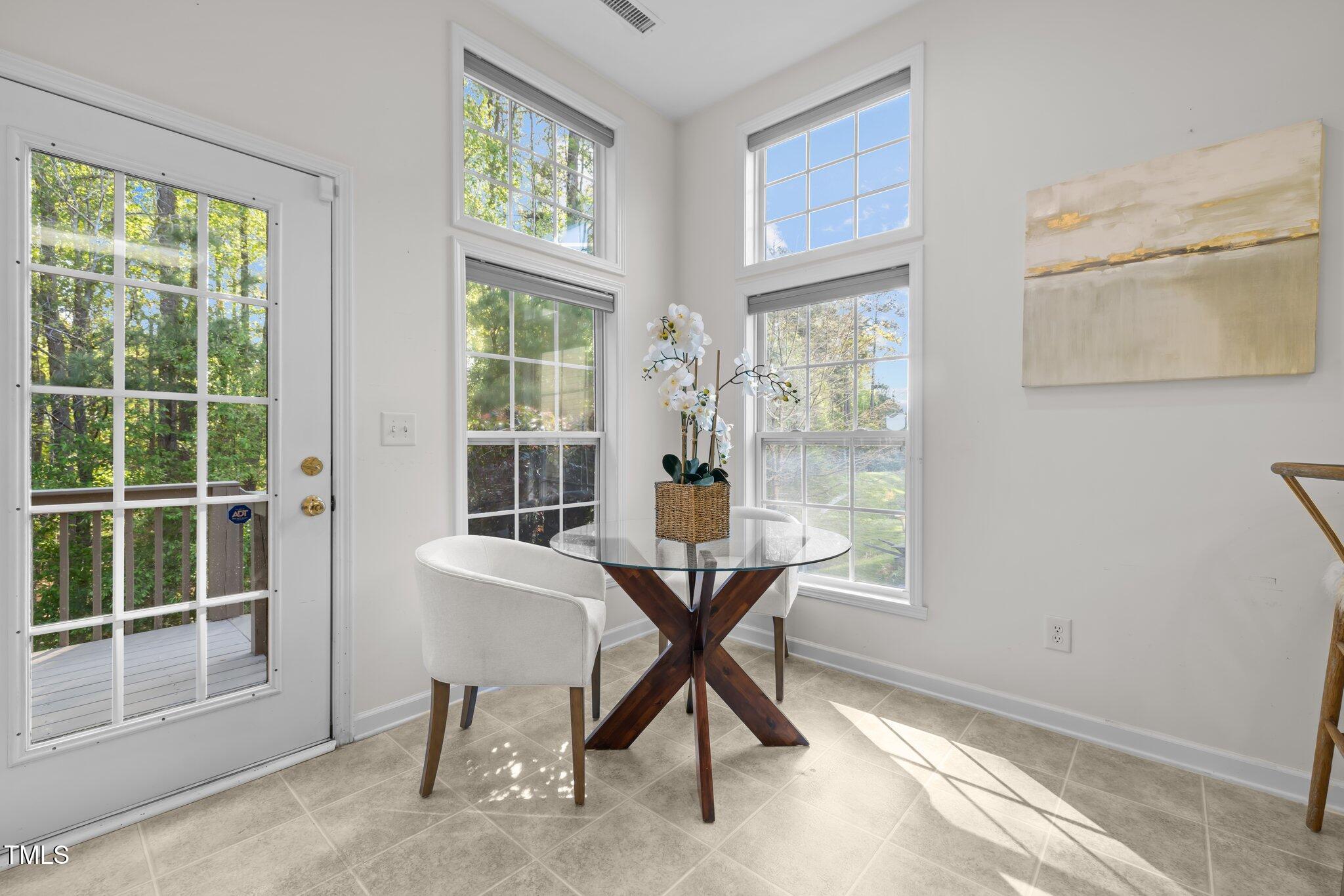 Bright dining nook with a glass-top table, white chairs, tall windows, and a glass-paneled deck door.