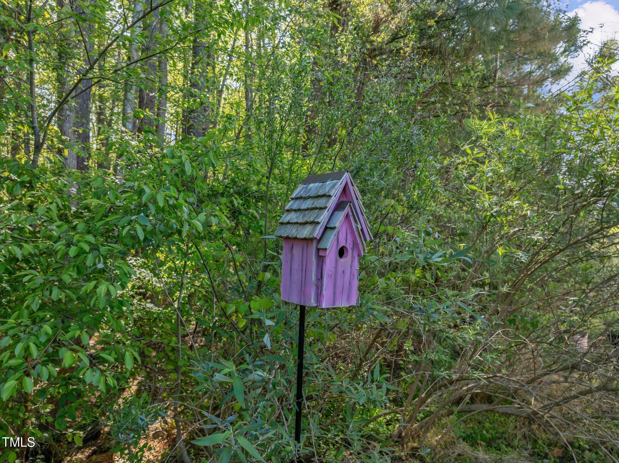 Purple wooden birdhouse with a shingled roof stands among leafy shrubs and trees.