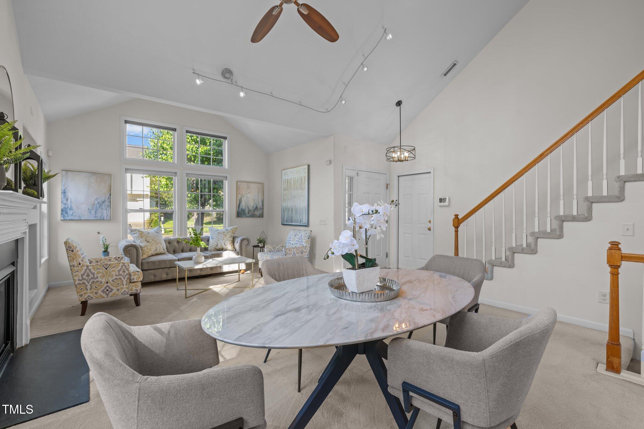 Furnished living and dining room with vaulted ceiling, tall windows, fireplace, and staircase.