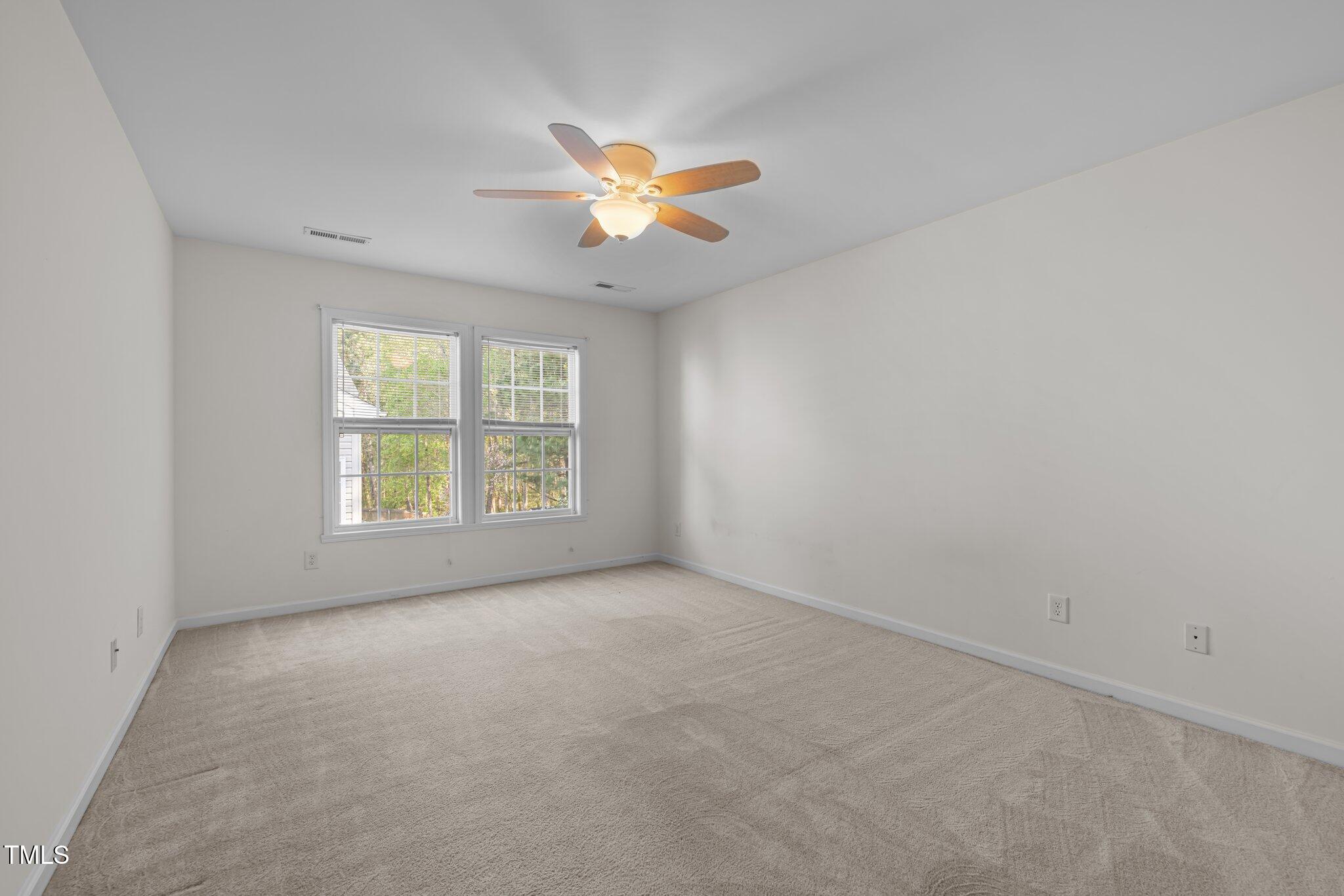 Empty bedroom with beige carpet, two windows, and a lit ceiling fan.