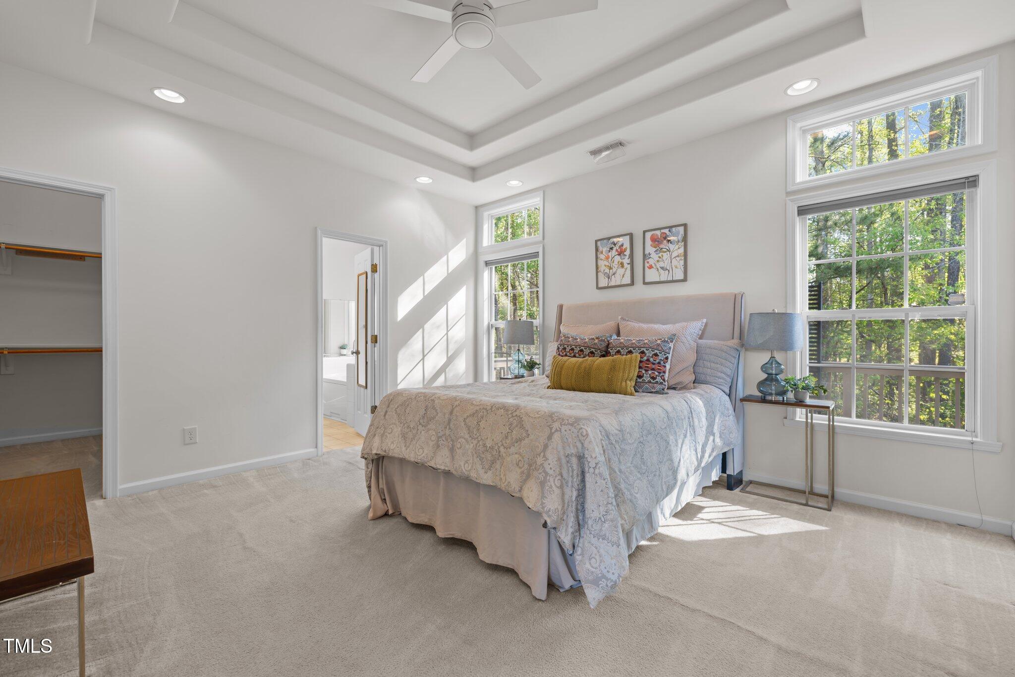 Furnished bedroom with a tray ceiling, large windows, and leafy trees outside.
