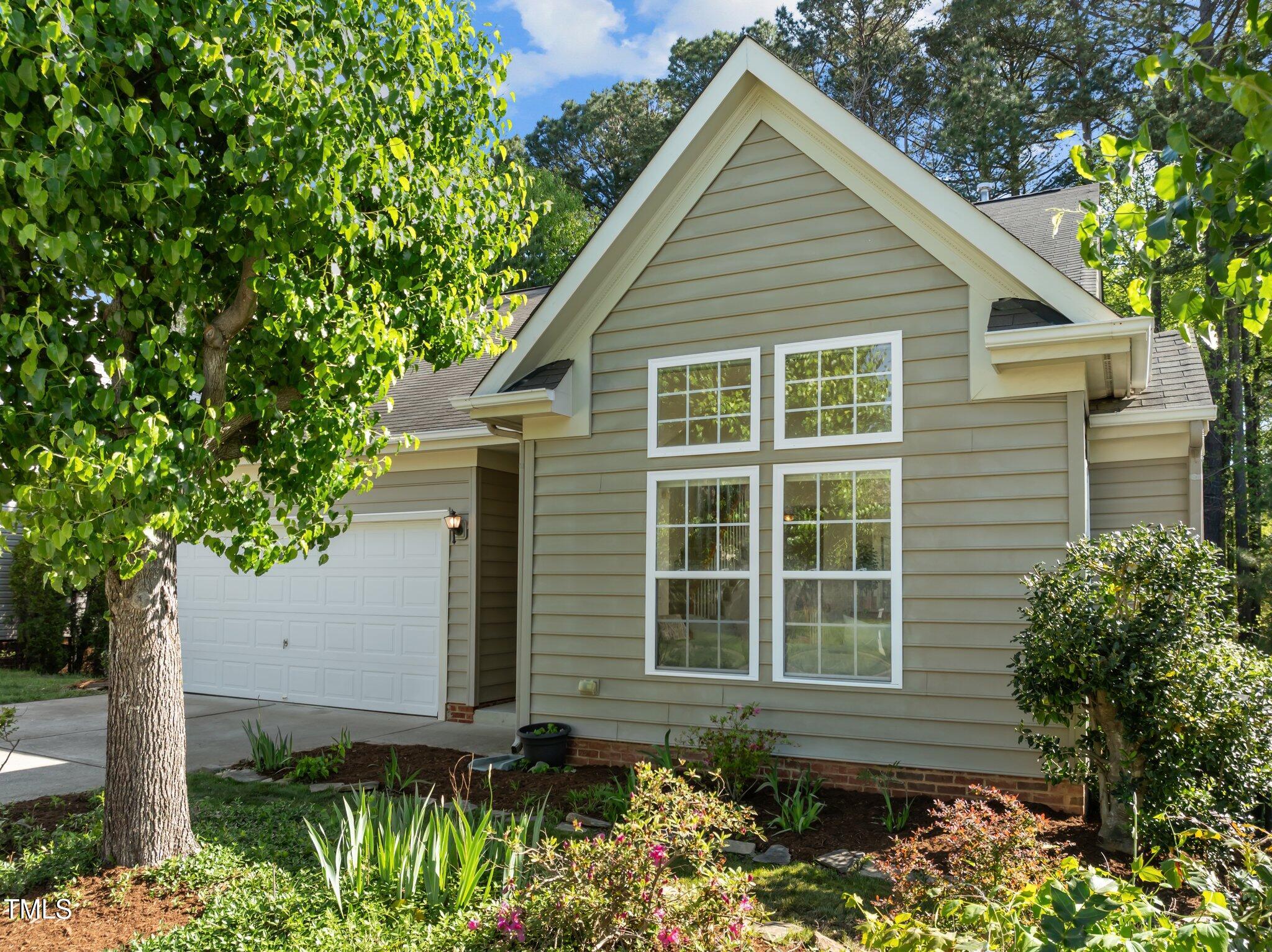 Two-story beige house with a steep gable, white-trimmed windows, and landscaped garden beds.