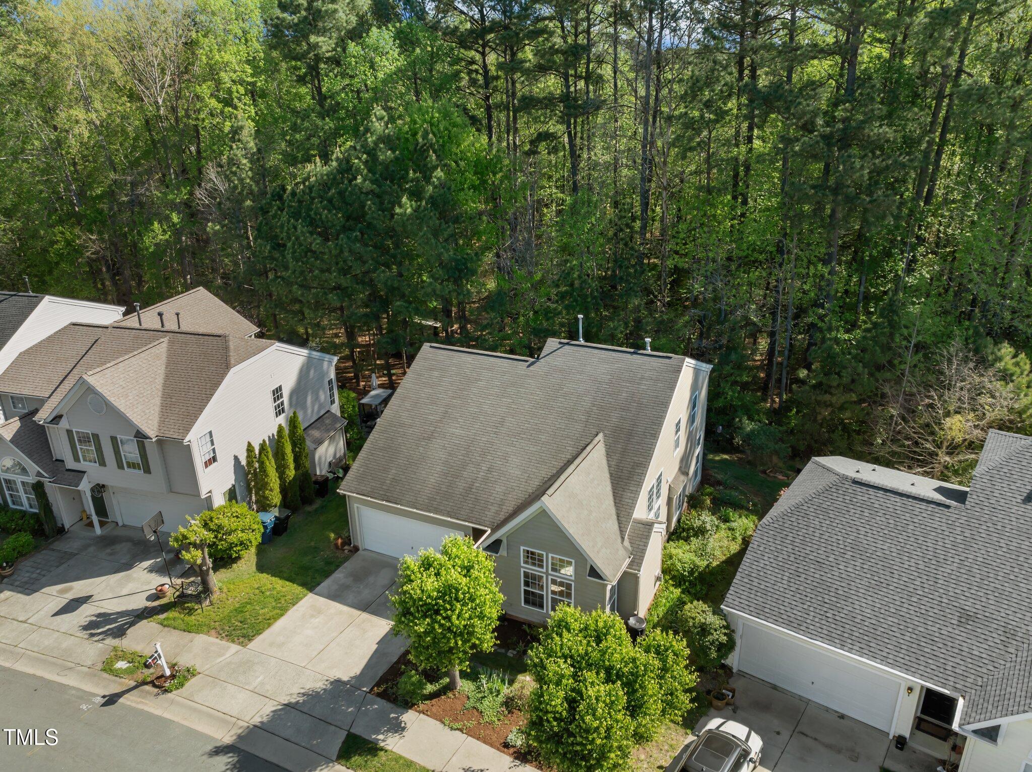 Aerial view of the home with a two-car garage, neighboring houses, and dense woods behind it.