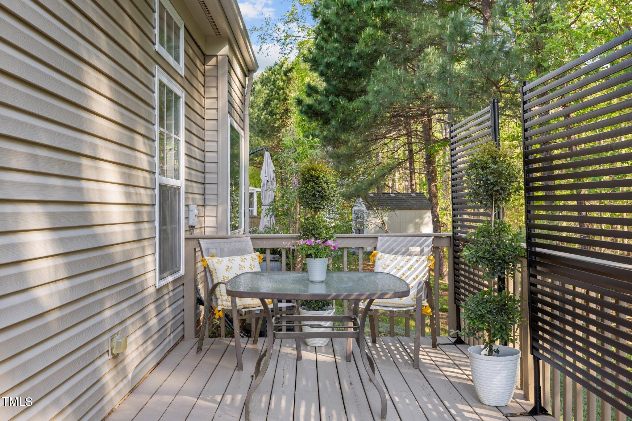 Outdoor deck with a glass-top table, cushioned chairs, potted flowers, and a slatted privacy screen.